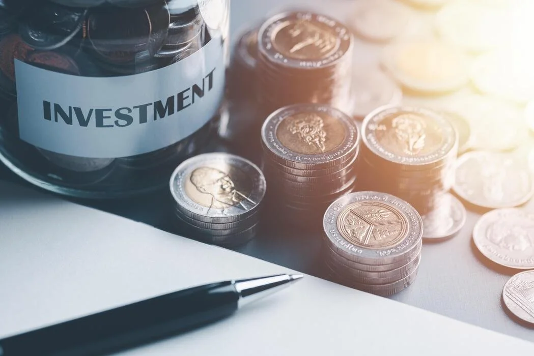 Coins stacked and scattered on a table next to a jar labeled 'Investment', with a pen and paper in the foreground.
