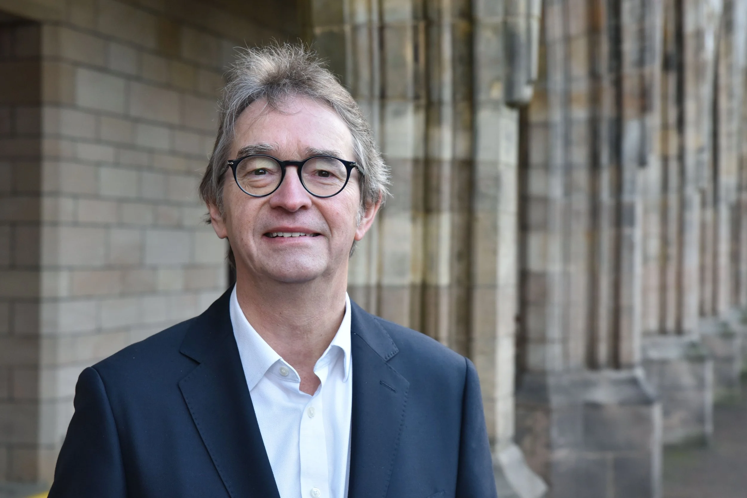 Portrait of a middle-aged man with gray hair and glasses, wearing a dark suit and white shirt, standing outdoors on a city street with a building in the background.
