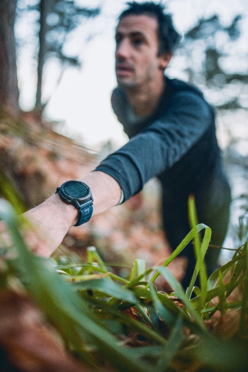 Kilian Jornet climbing a steep trail, with greenery and trees in the background. Kilian Jornet is wearing a Coros watch and a long sleeve shirt.