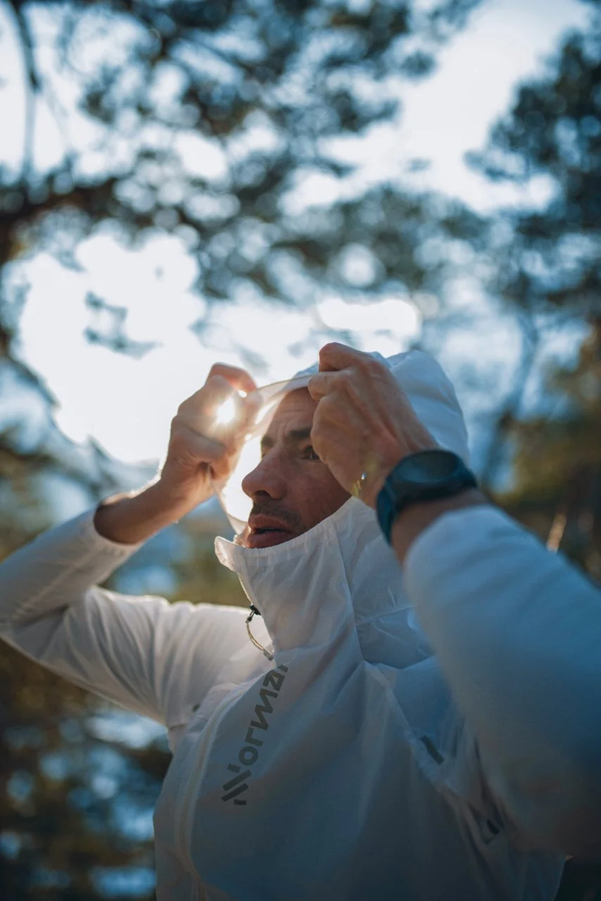 Kilian Jornet in a white NNormal jacket adjusting the hood, with sunlight filtering through trees in the background.