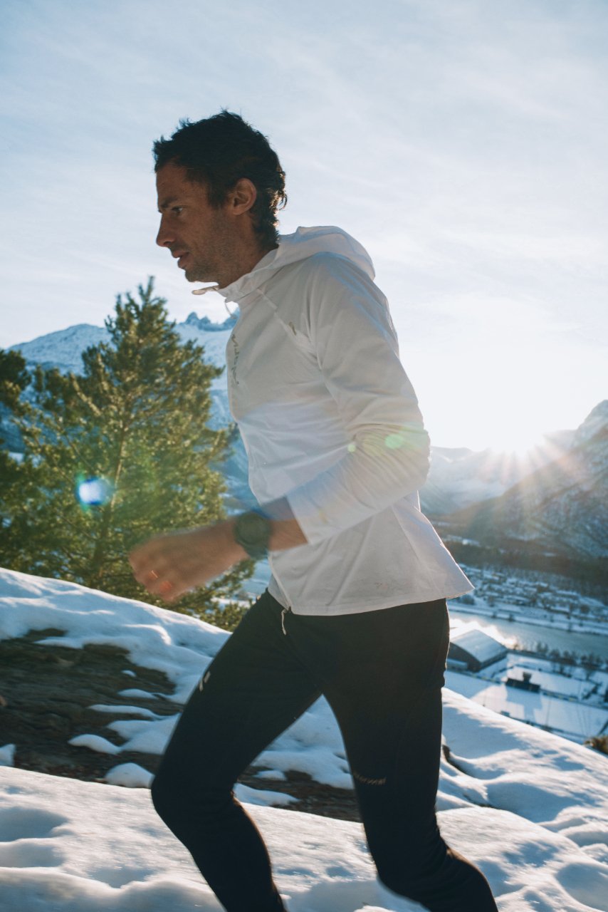 Kilian Jornet running outdoors in a winter landscape with snow, trees, mountains, and a clear blue sky, wearing a white NNormal jacket and black pants.