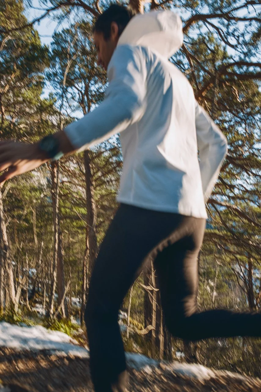 Kilian Jornet running outdoors through a wooded area with trees and snow on the ground, wearing a white NNormal jacket and black pants.
