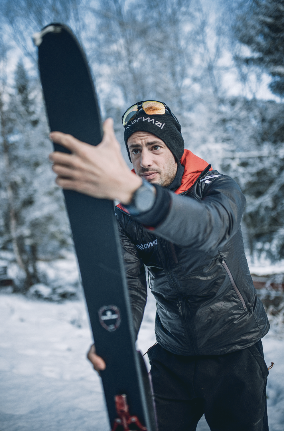 Kilian Jornet outdoors in a snowy forest mixes up ski equipment, wearing a black jacket, a beanie, and sunglasses on his forehead.