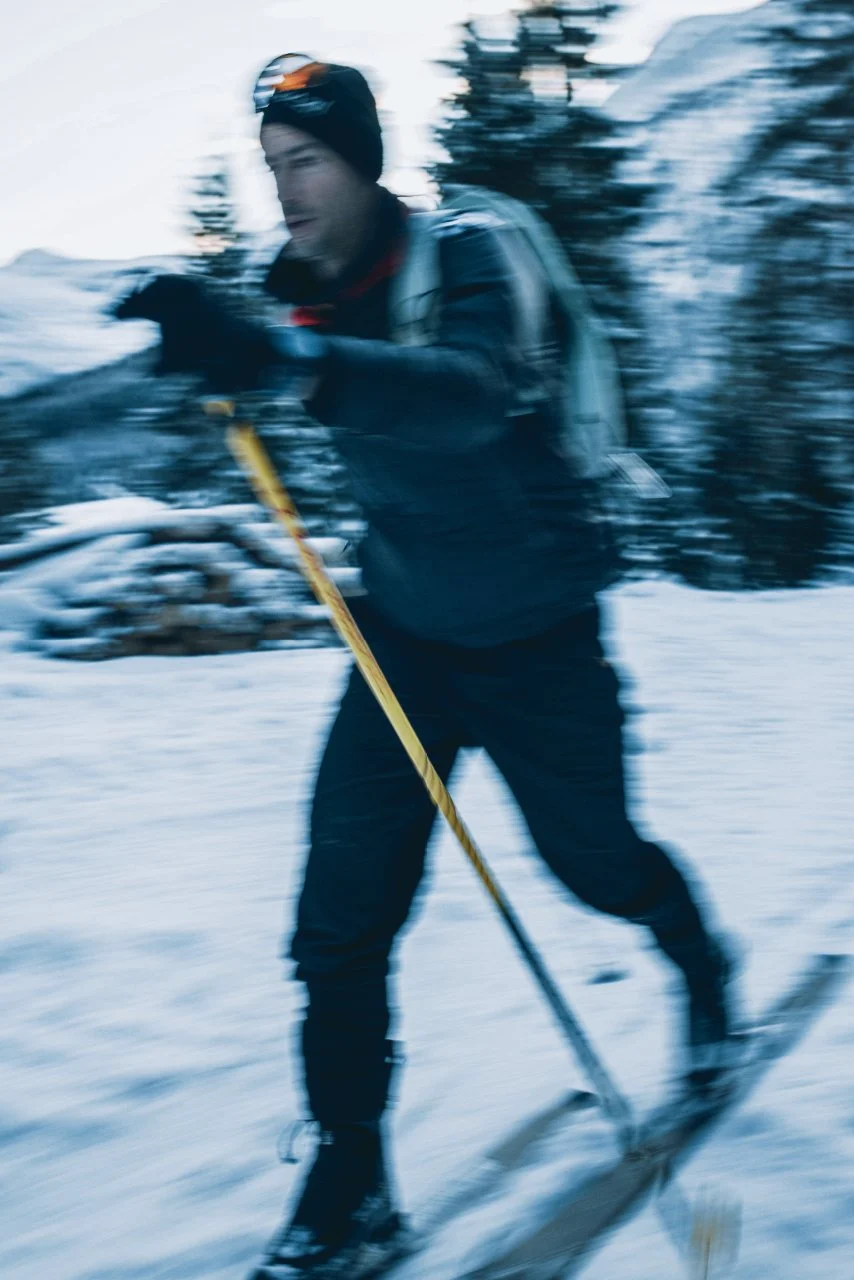 Kilian Jornet cross-country skiing in snowy woods, wearing winter gear including gloves and a hat with goggles, holding ski poles, with a motion-blurred background indicating movement.