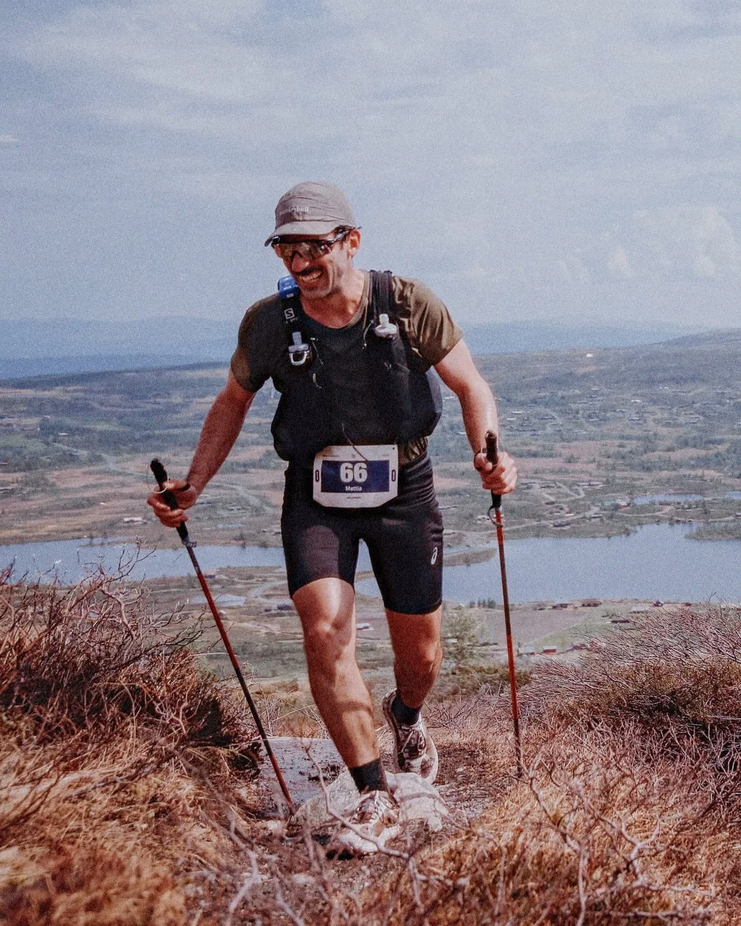 Mattia Lullini running uphill in a trail race, dressed in athletic gear with a race bib number 66 and trees and a lake in the background.