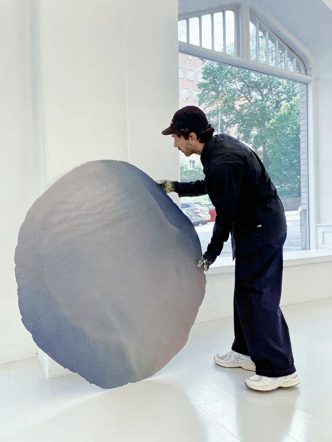 Mattia Lullini wearing a black jacket, black cap, and white sneakers is working on a large, circular art piece indoors at NEVVEN front of a large window, with trees and buildings visible outside.