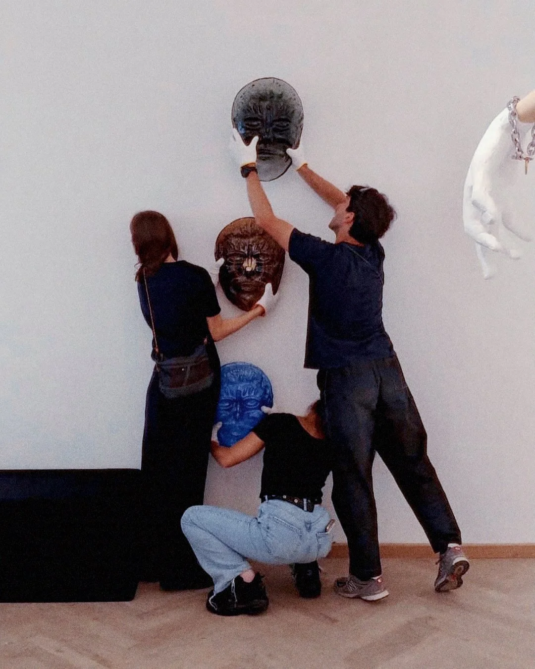 Three people hanging masks on a white wall in an NEVVEN. Two adults are attaching masks while a child holds a blue mask.