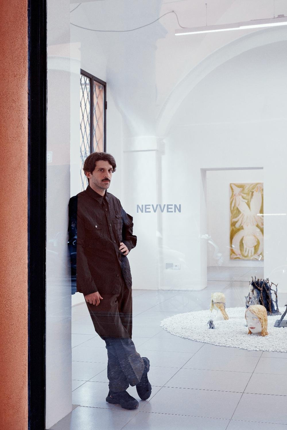 Mattia Lullini dark hair and mustache wearing a black shirt and dark pants leaning against glass at NEVVEN with sculptures and paintings visible inside.