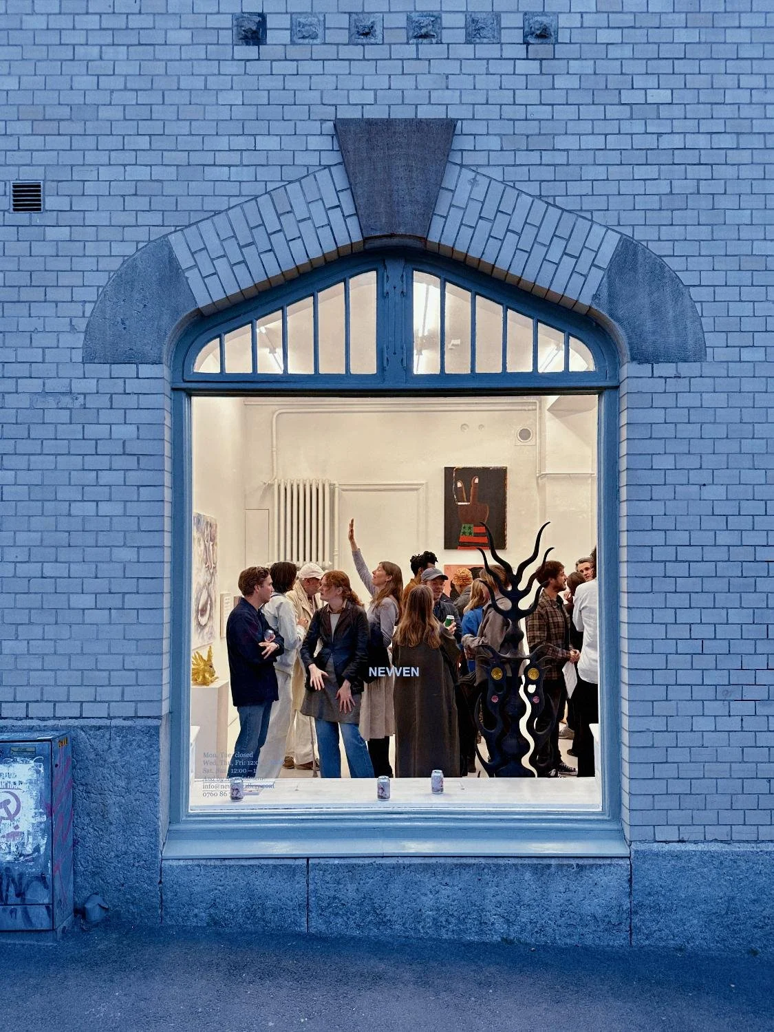 People inside an art gallery, viewed through a large window, with artwork and sculptures visible, and a sign that says 'NEVVEN'.