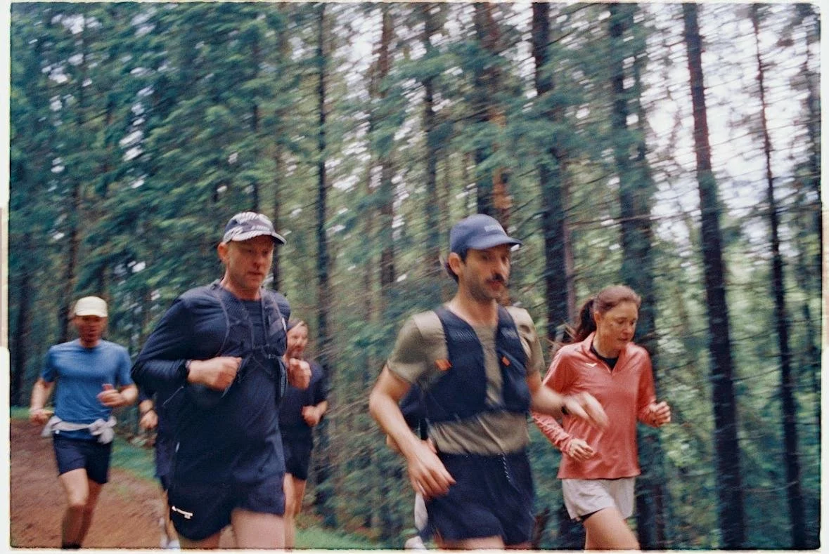 Group of five people trail running through a forested area with tall trees.