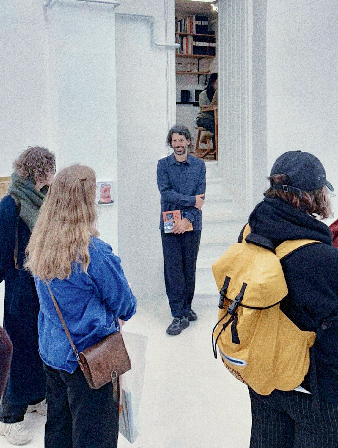 A man stands in an art gallery or museum, holding a book, and smiling while a group of people listens to him. The setting has white walls and a staircase in the background.