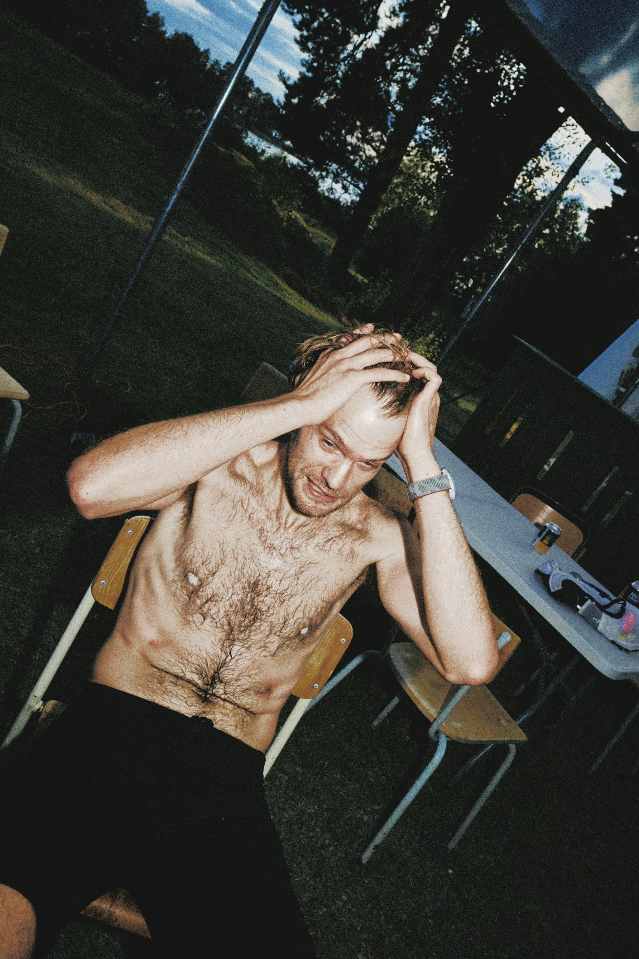 Sörmland 100 A shirtless man with messy hair sitting outdoors, holding his head in frustration or stress, at twilight or early evening with trees and outdoor furniture in the background.