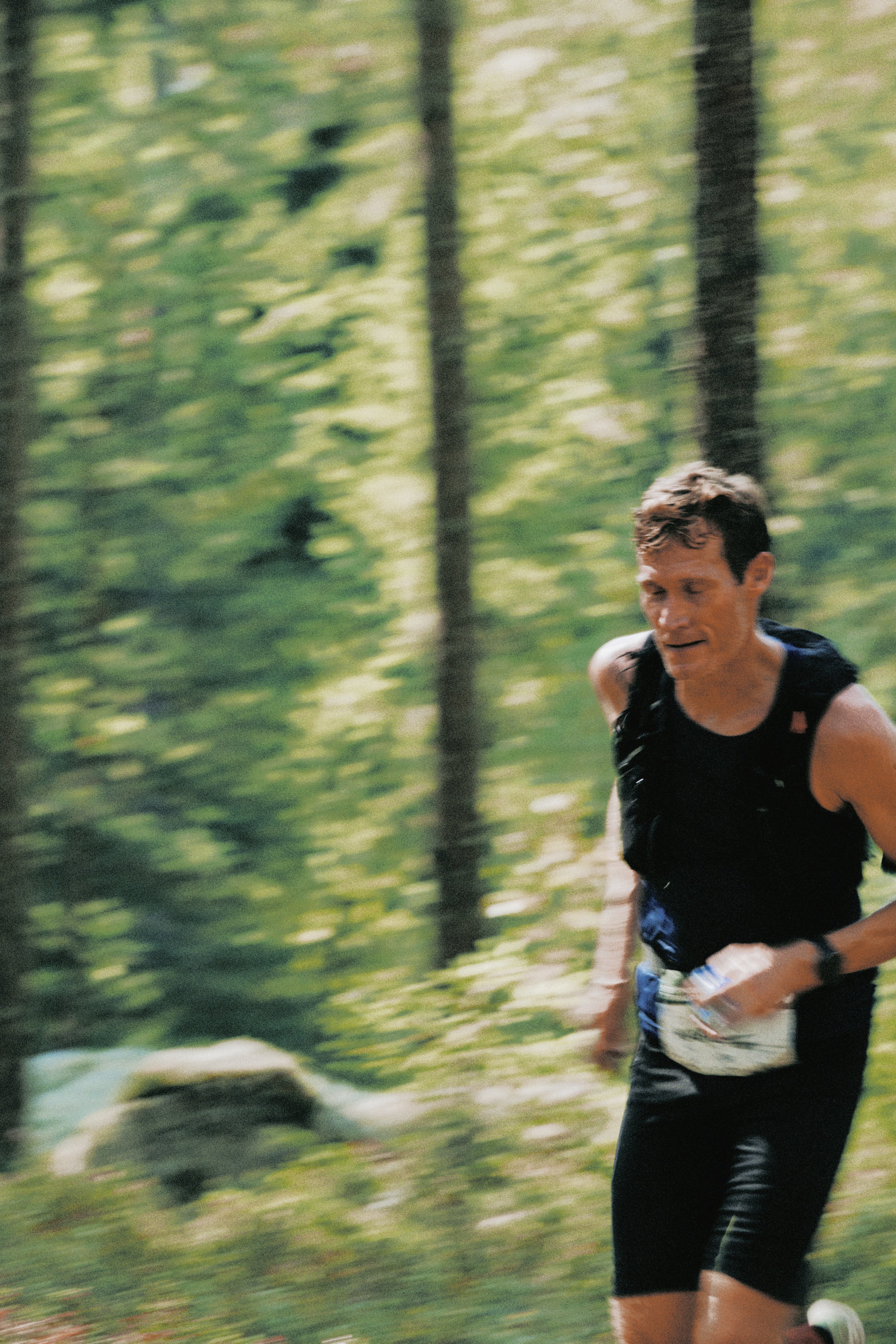 Sörmland 100 A man running through a green forest during a race, holding a water bottle, wearing a black sleeveless shirt and black shorts.