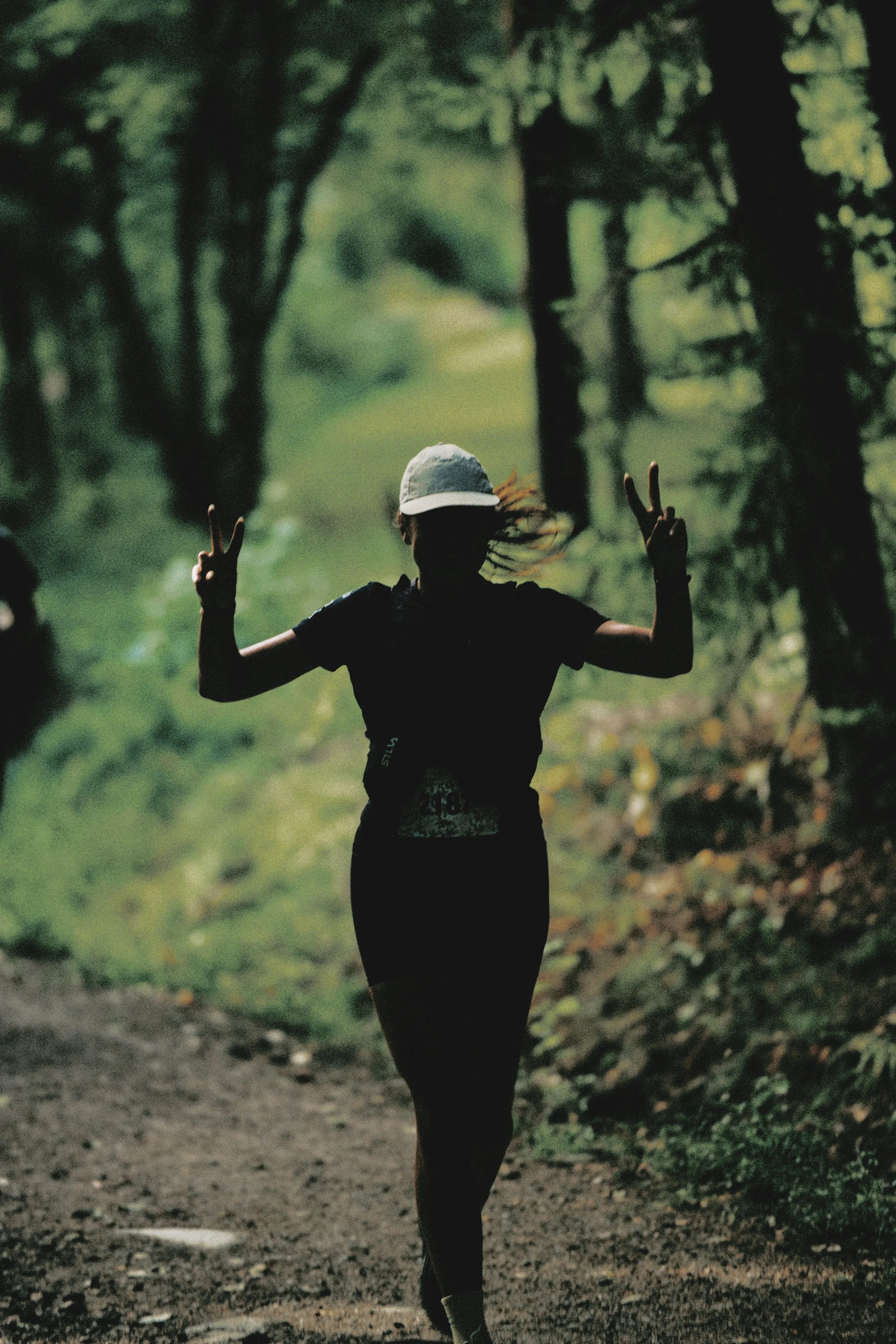 Sörmland 100 Silhouette of a woman walking on a trail in a wooded area, making peace signs with both hands.