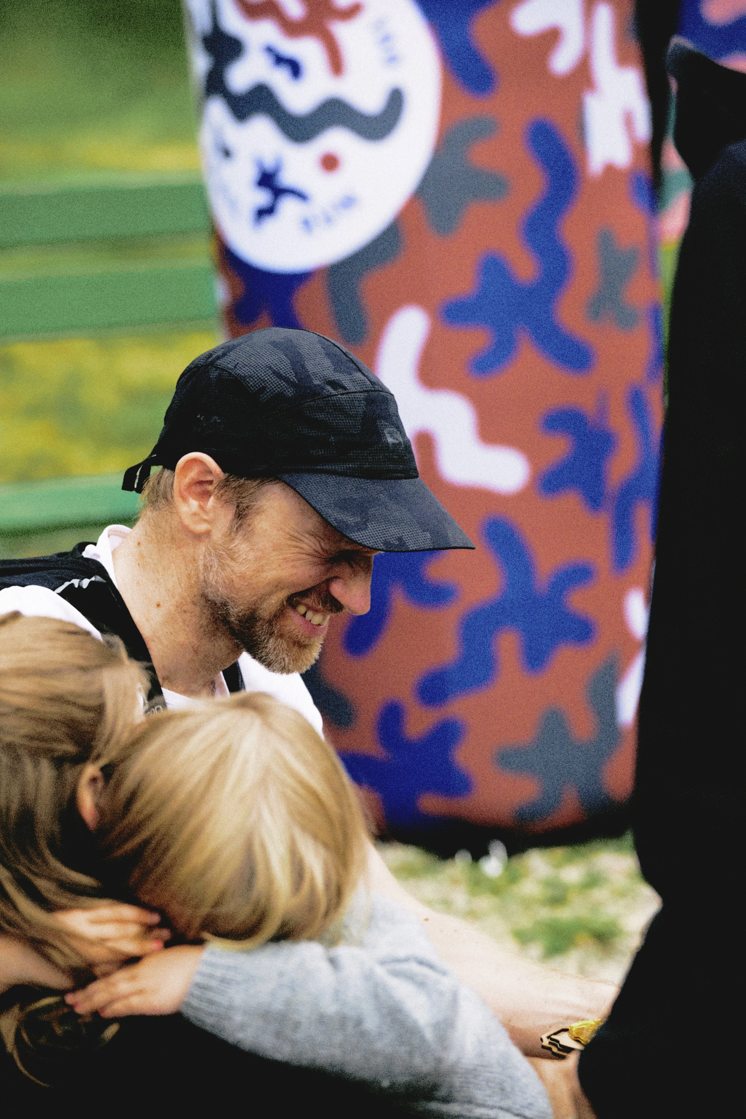 Sörmland 100 A man with a beard and black cap laughing with two children, one with blonde hair, on an outdoor bench or table, with colorful abstract art in the background.