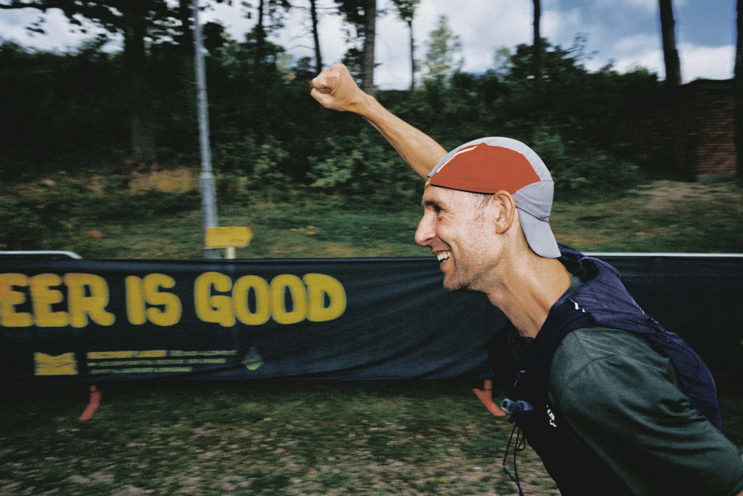 Sörmland 100 Man running outdoors with a red and gray cap, smiling, and a banner in the background that reads 'BEER IS GOOD'.