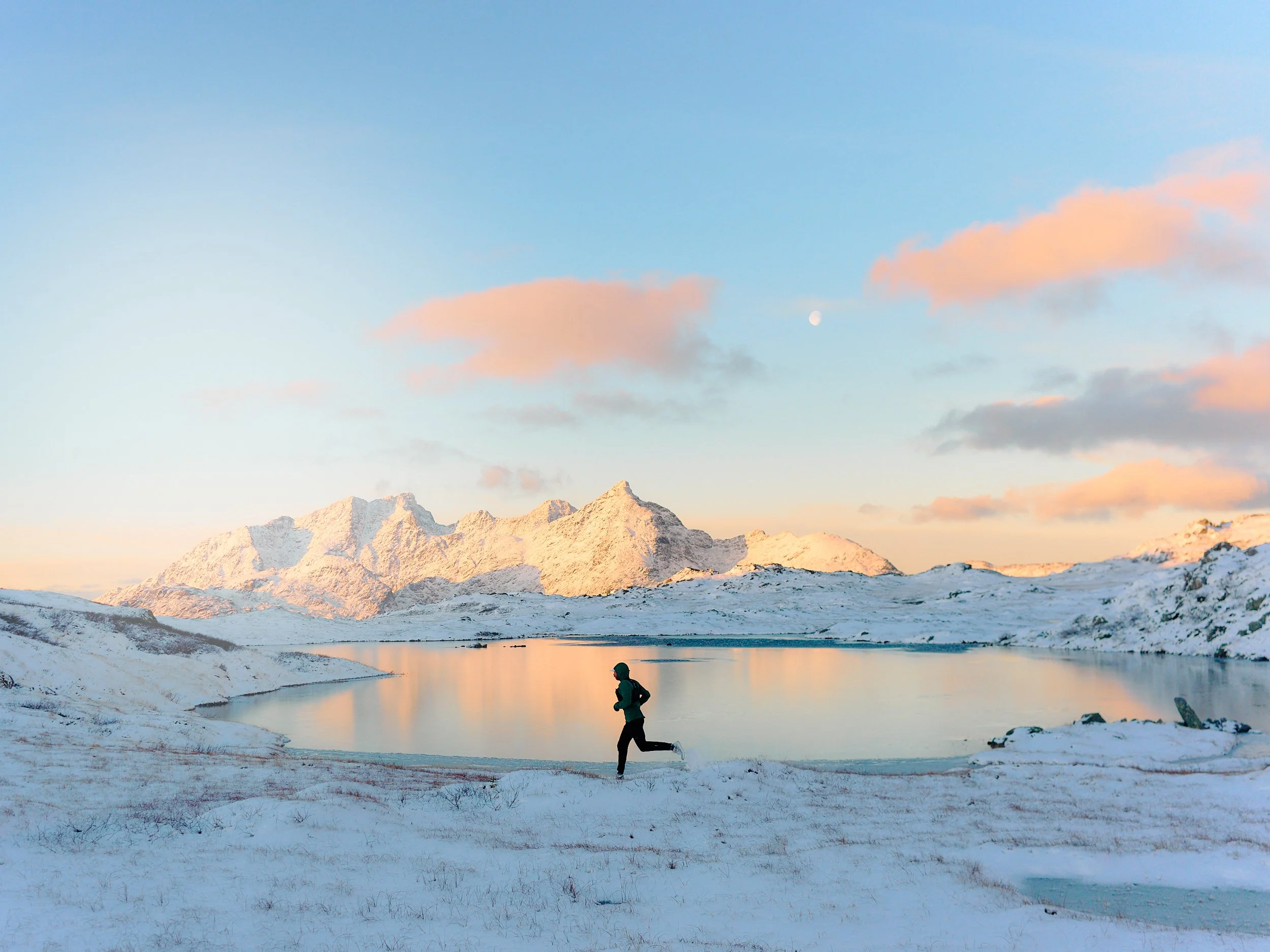 A person running along a snowy lakeshore with snow-covered mountains in the background under a partly cloudy sky.