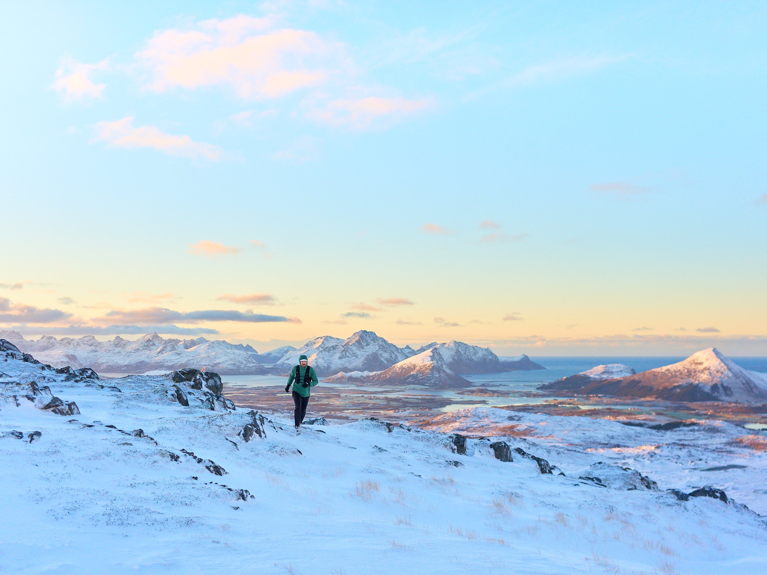 A person hiking on a snow-covered mountain ridge with a view of distant snow-capped mountains, water, and a partly cloudy sky during sunrise or sunset.
