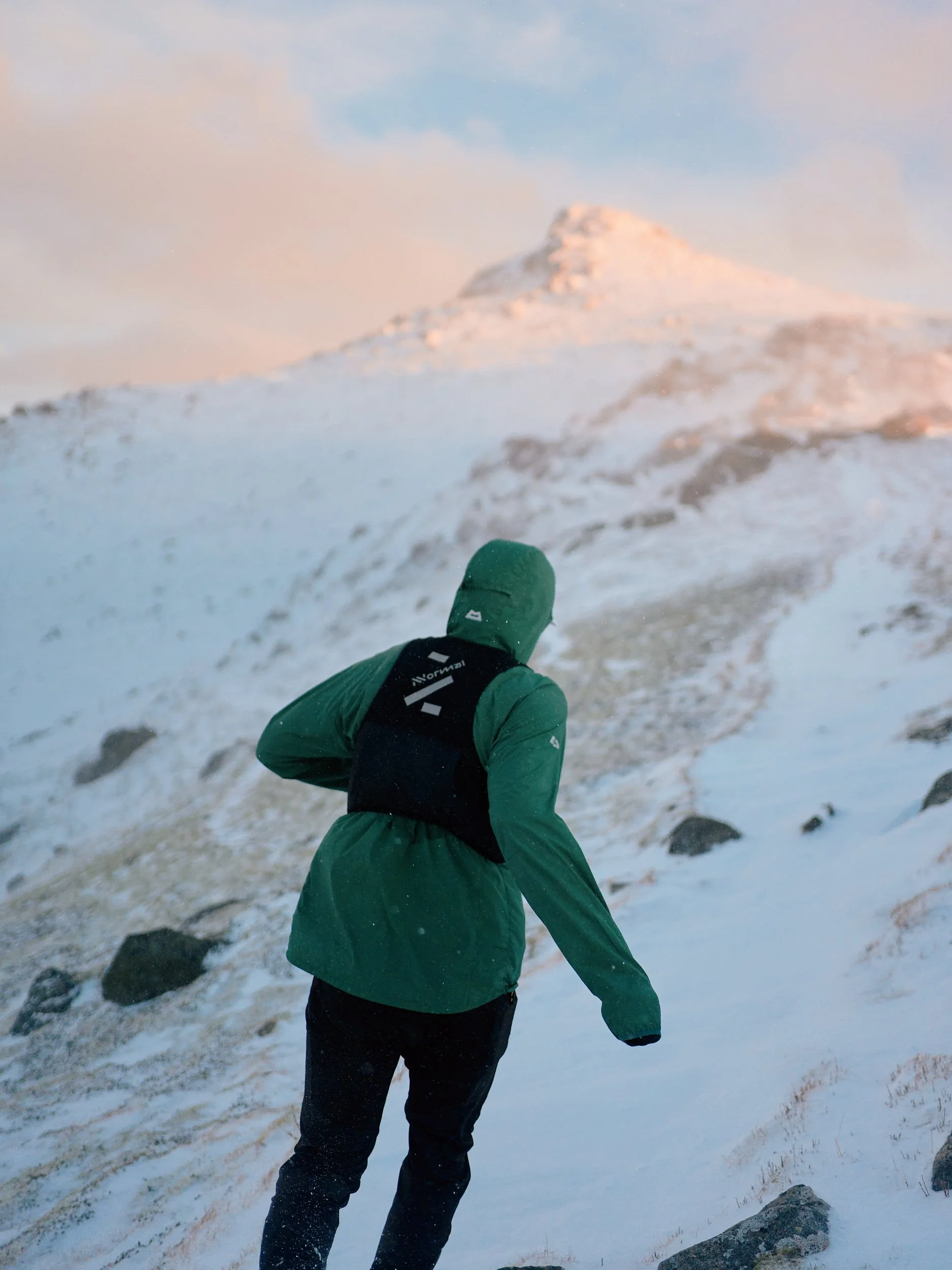 Person dressed in green outdoor gear hiking or climbing on snowy mountain terrain during sunrise or sunset.