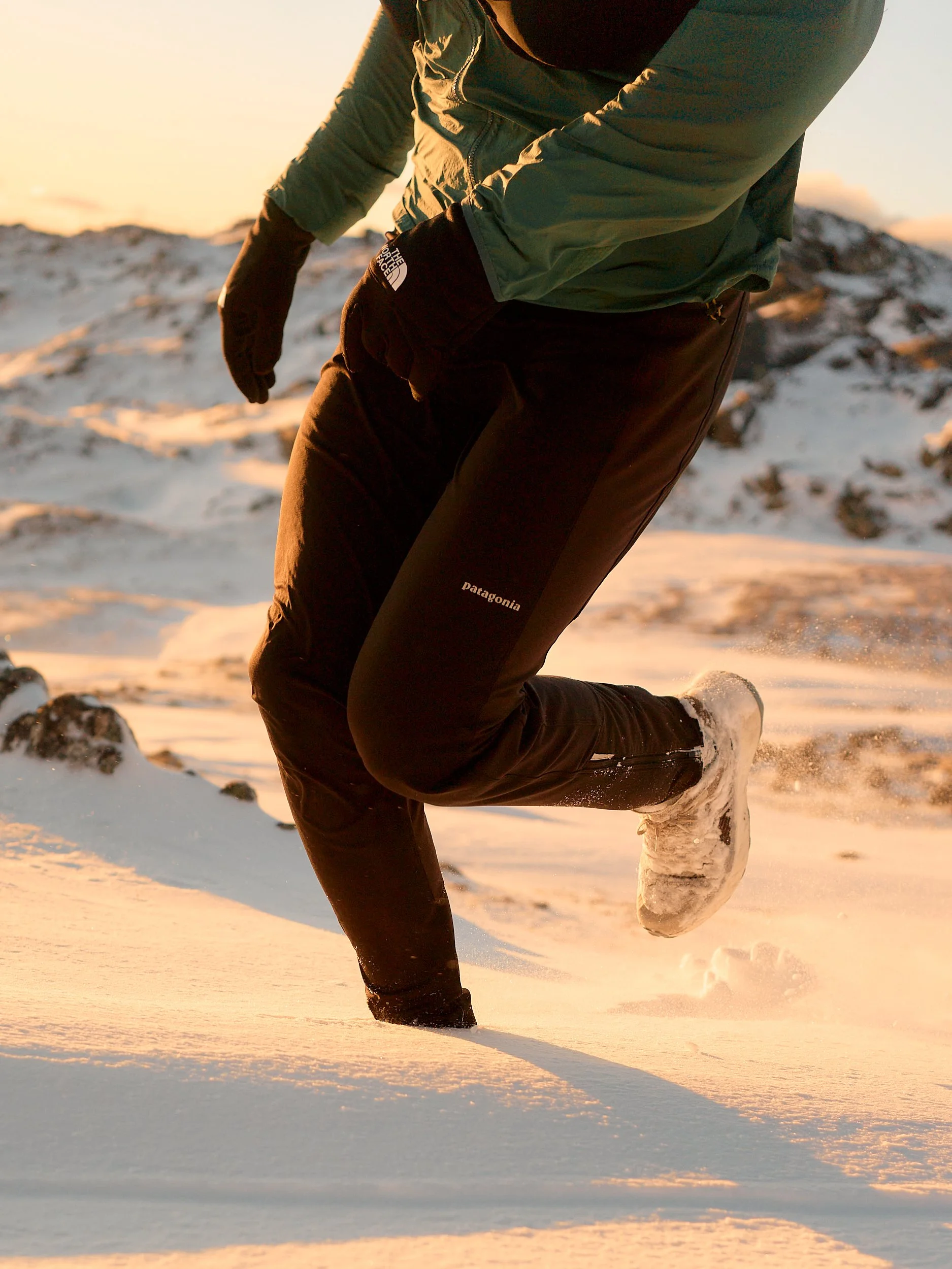 A person running in the snow wearing black Patagonia pants, a green jacket, black gloves, and white shoes, with snow and rocks on the ground at sunset.