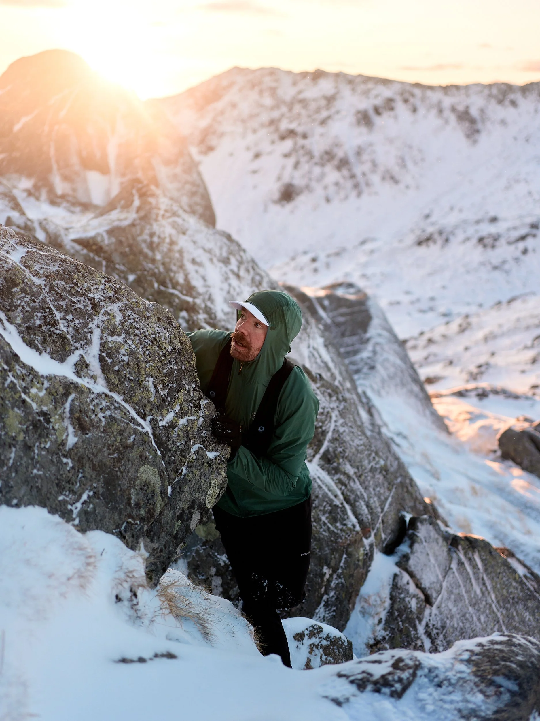 A man wearing a green and black jacket, black gloves, and a white cap is climbing a snowy and rocky mountain in the early morning or late afternoon, with the sun shining behind the peaks.