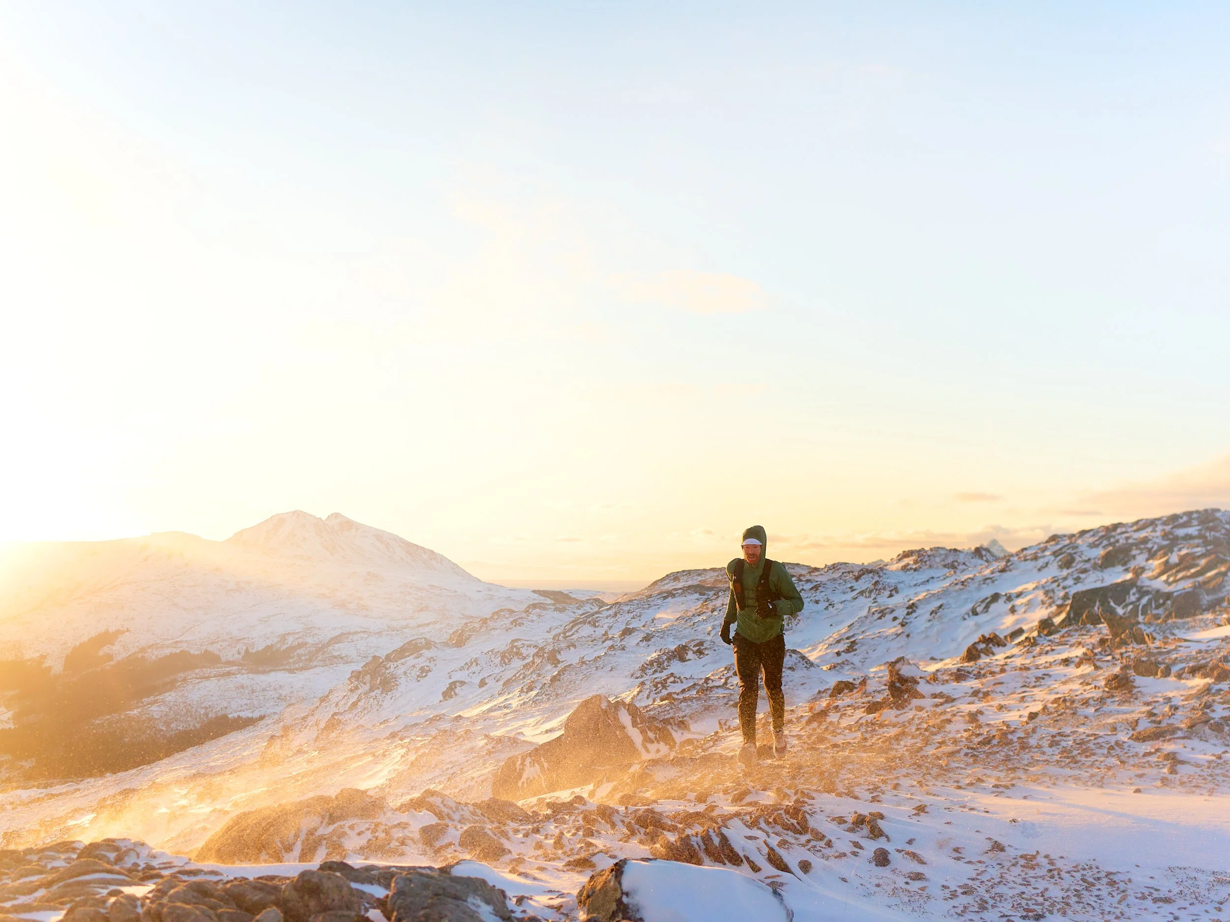 A man hiking in snow-covered mountainous landscape at sunrise or sunset.