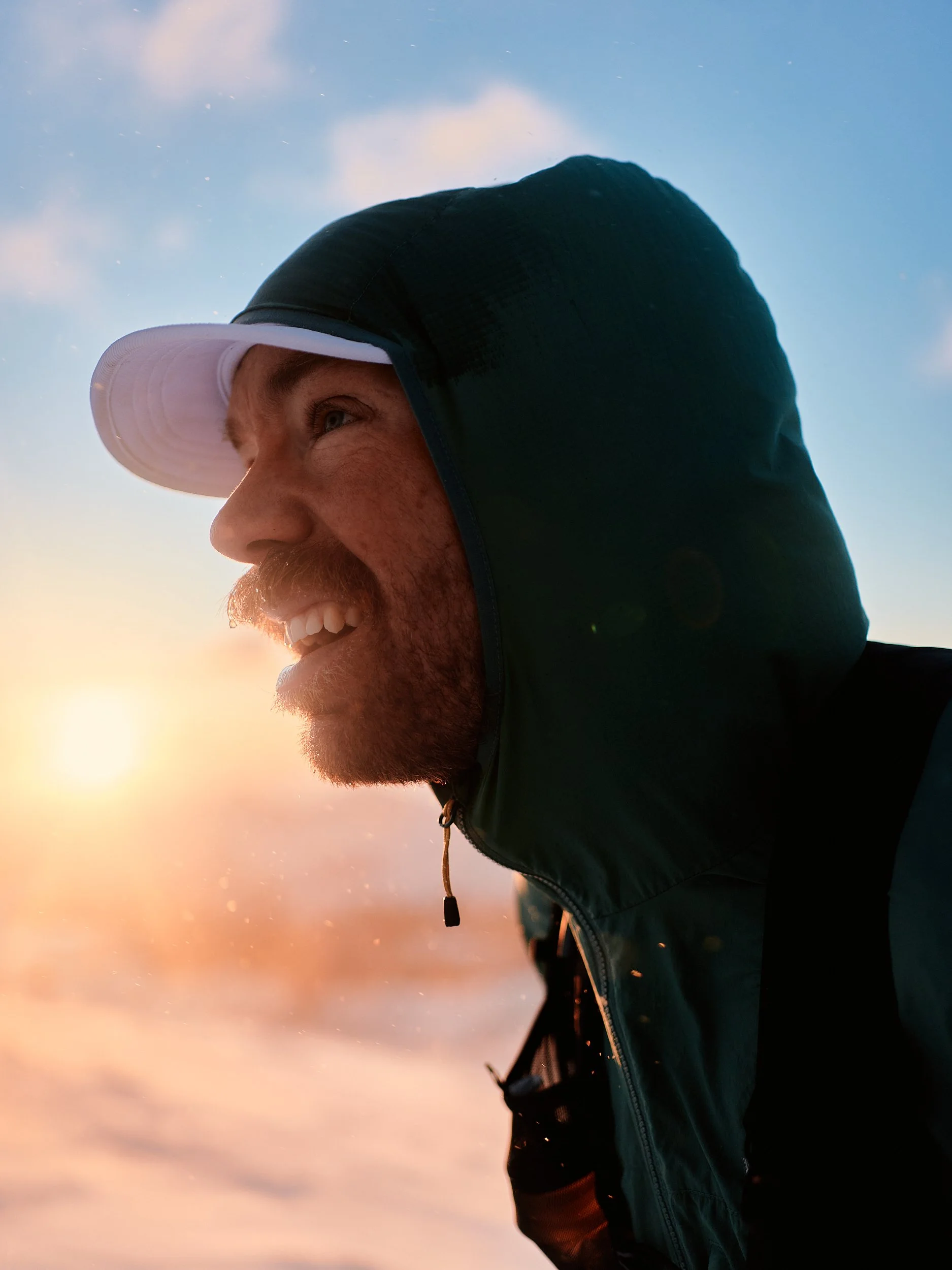 A smiling man wearing a jacket with a hood and a white cap, outdoor during sunrise or sunset.