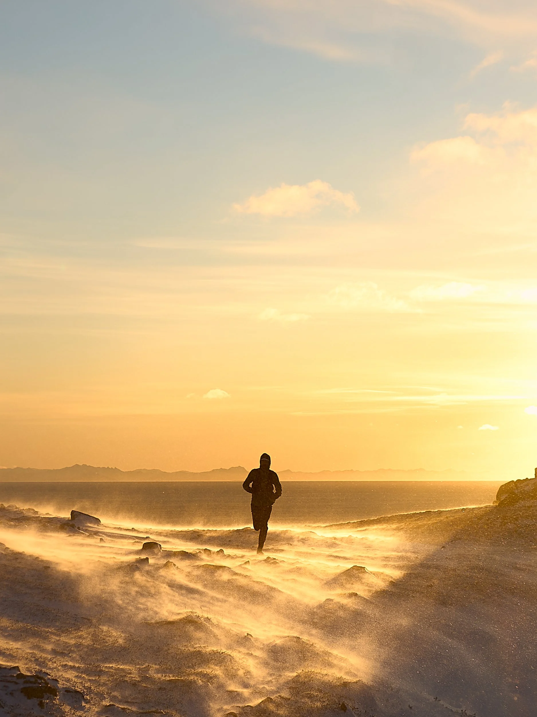 Andreas Køhn running on a windy mountain in Lofoten during sunset with a distant mountain range on the horizon and a partly cloudy sky.