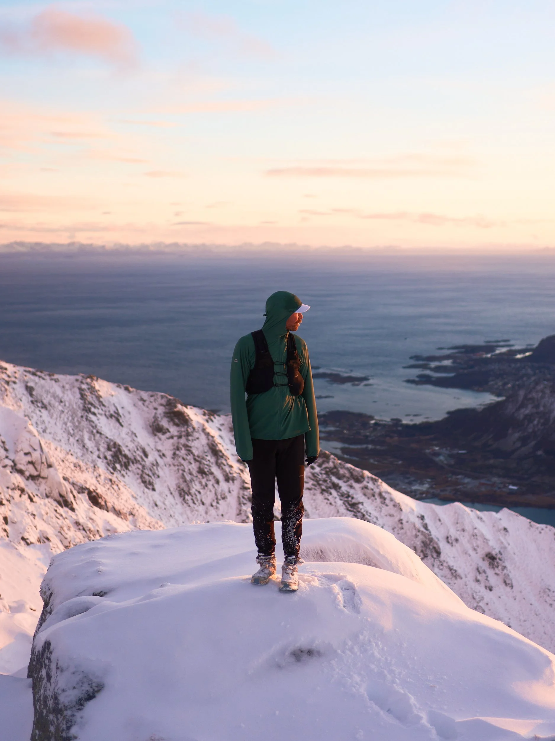 Person dressed in outdoor gear standing on snow-covered mountain peak during sunset, with a view of the ocean and distant islands.