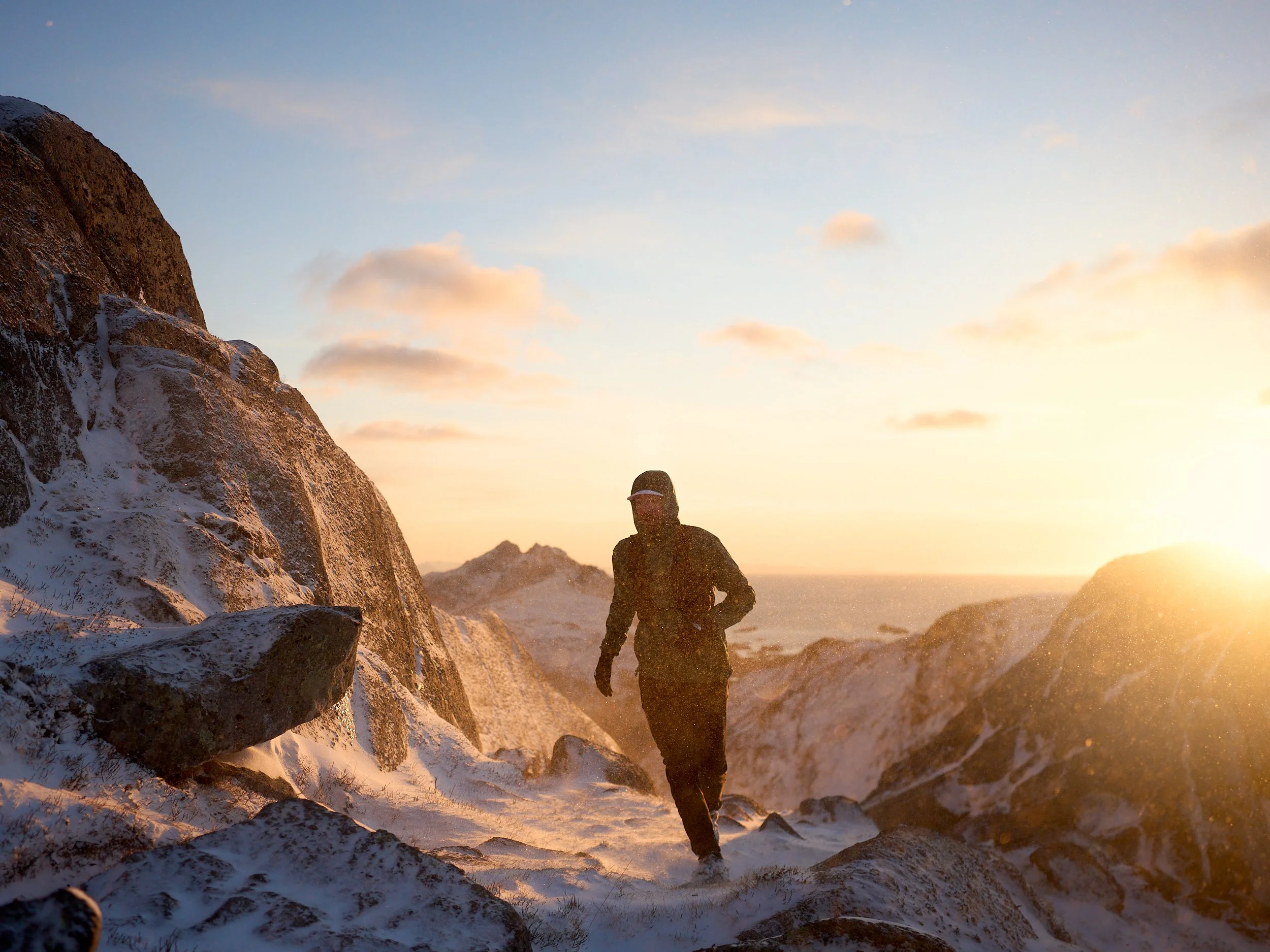 Person dressed in black running on snow-covered rocky terrain at sunset with mountains in the background.
