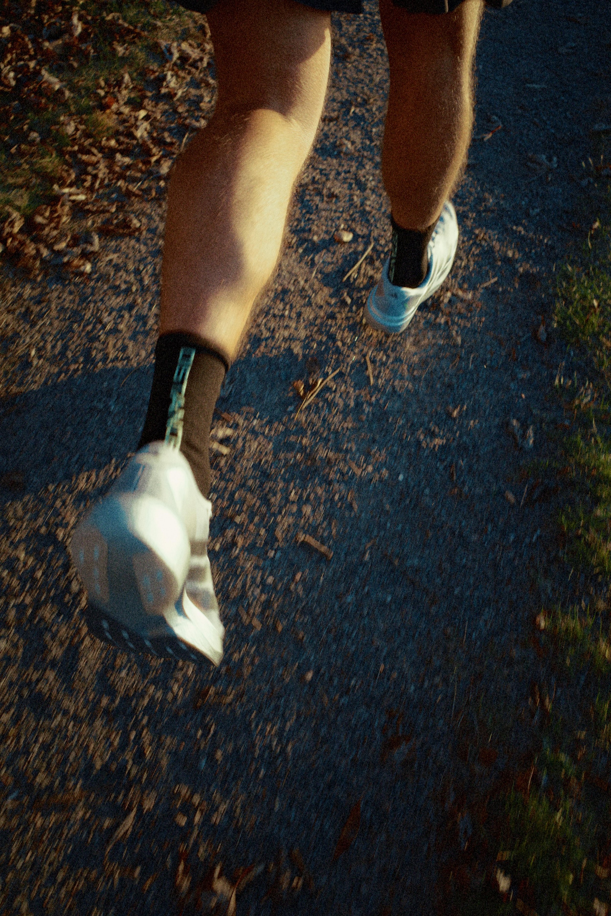 Close-up of a person's legs and feet running on a dirt trail during daytime, wearing white running shoes and dark socks.