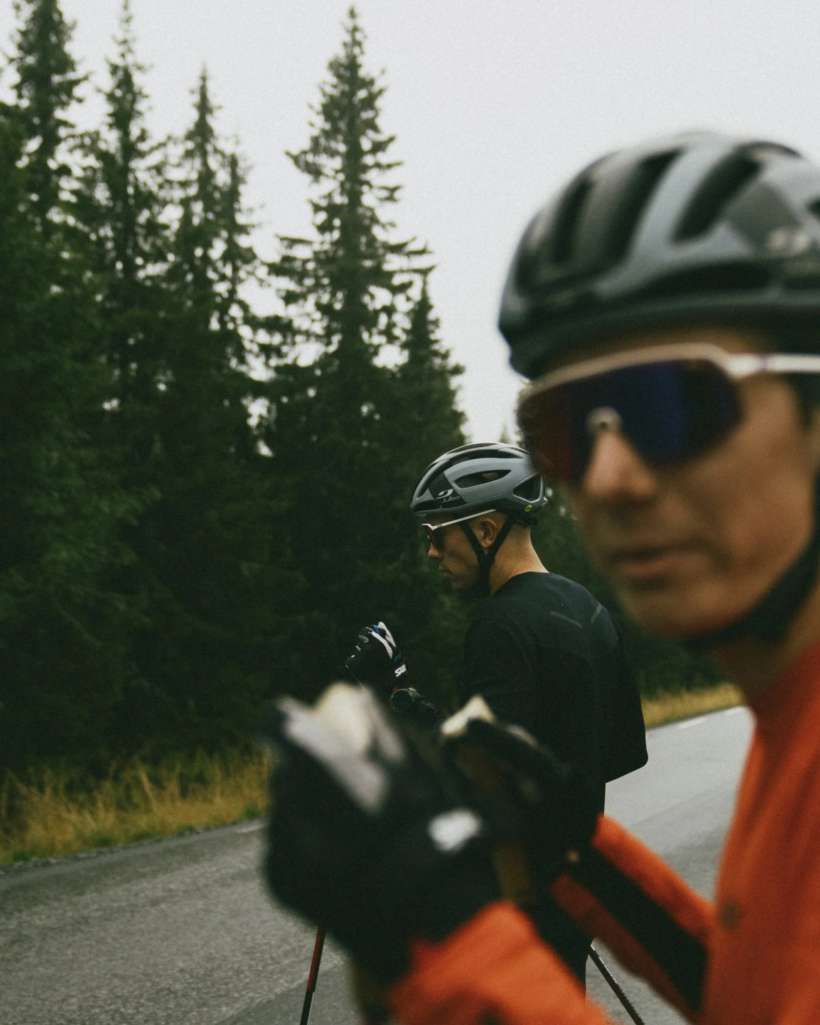 Two men Nordic skiing wearing helmets and sunglasses standing on a road with a background of trees and an overcast sky.