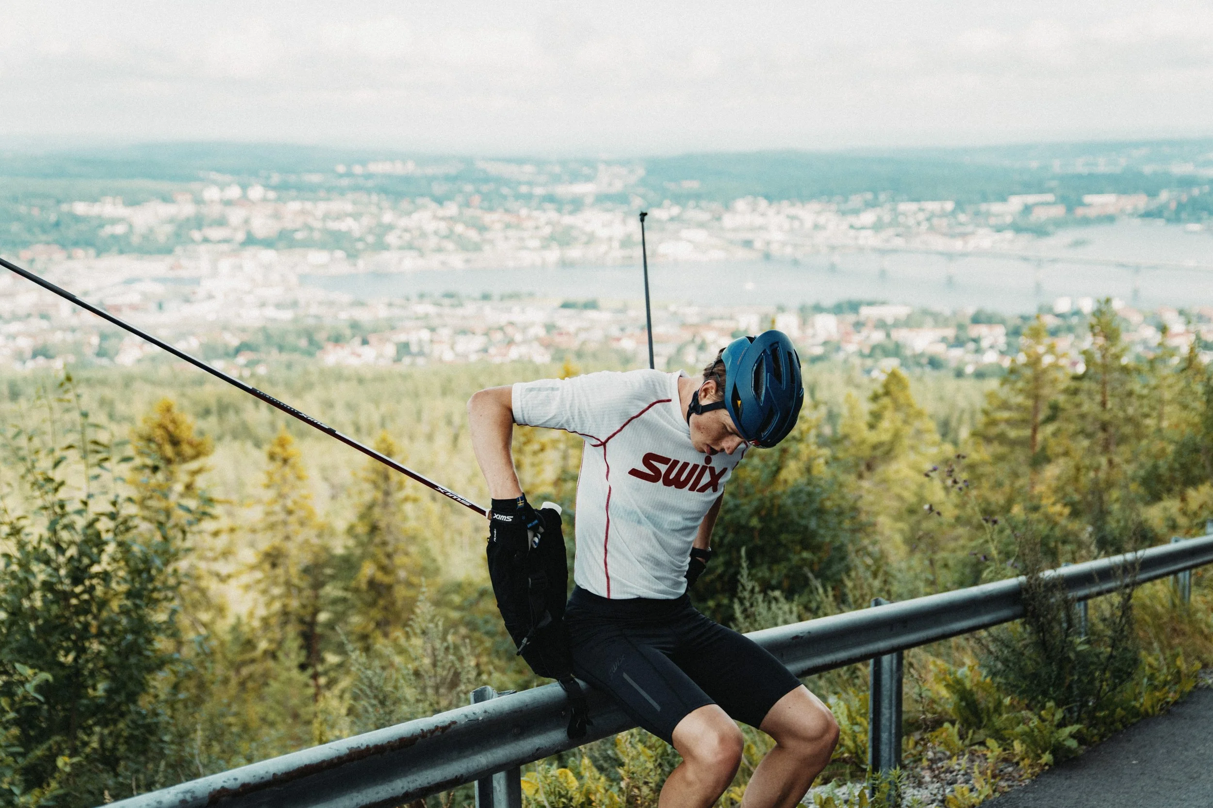 William Poromaa wearing Swix sports gear, including a helmet and gloves, sitting on a guardrail with a scenic view of a city and river in the background. Nordic roller ski training.