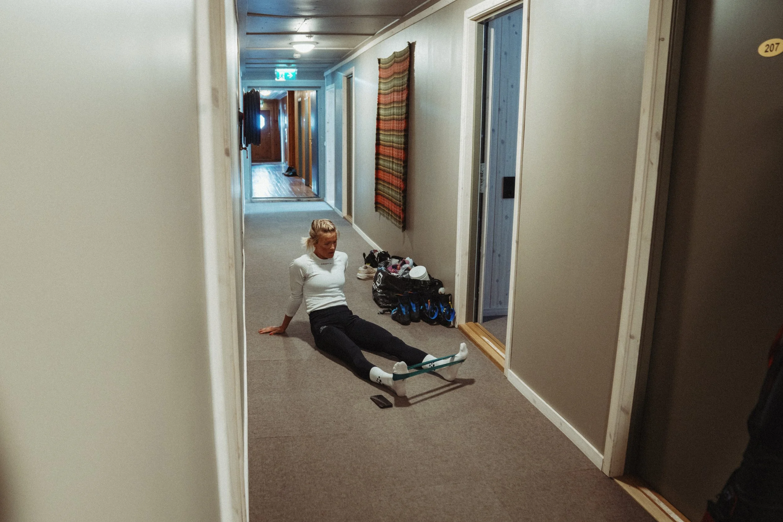 Frida Karlsson sitting on a hallway floor with a resistance band around her legs, leaning back on her hands, near a pile of shoes, bags, and personal items.