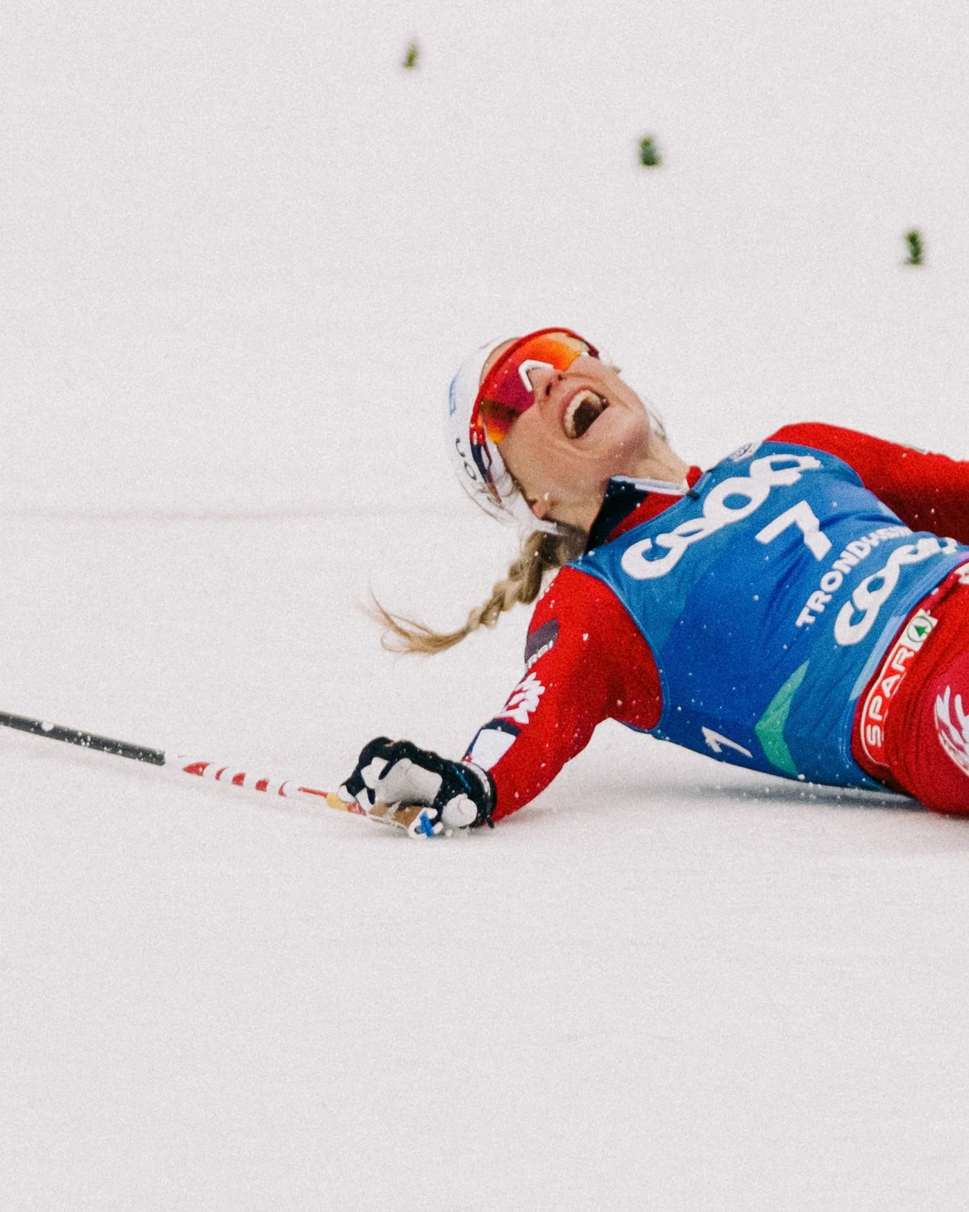 Therese Johaug lying on the snow with a big smile, wearing a blue bib, red and white gear, and sunglasses, during a skiing event, Trondheim 2025.
