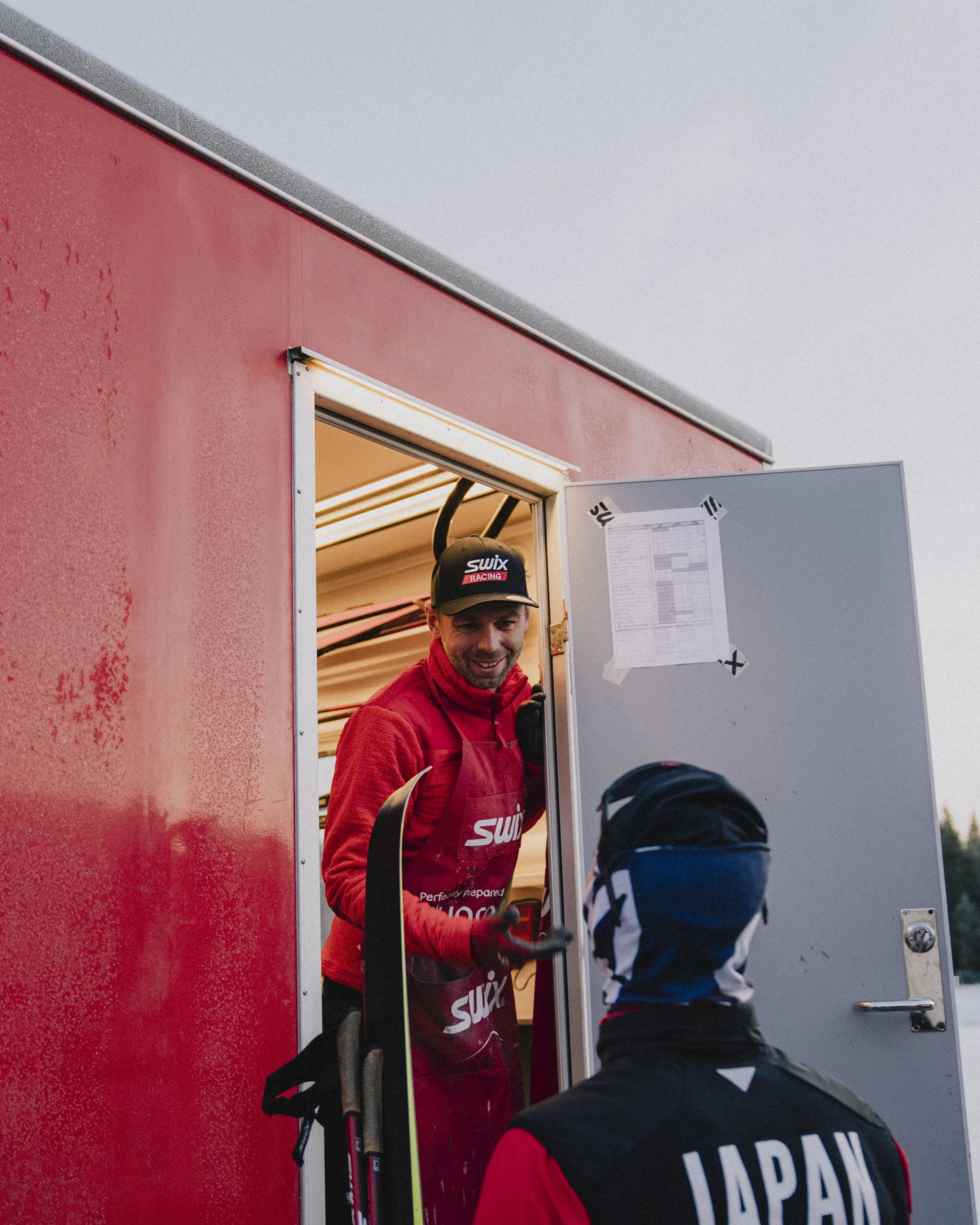SWIX racing athletes exchanging smiles at a red race event booth, one wearing a black cap and red outfit, and the other with a blue bandana and black jacket with 'JAPAN' written on the back.