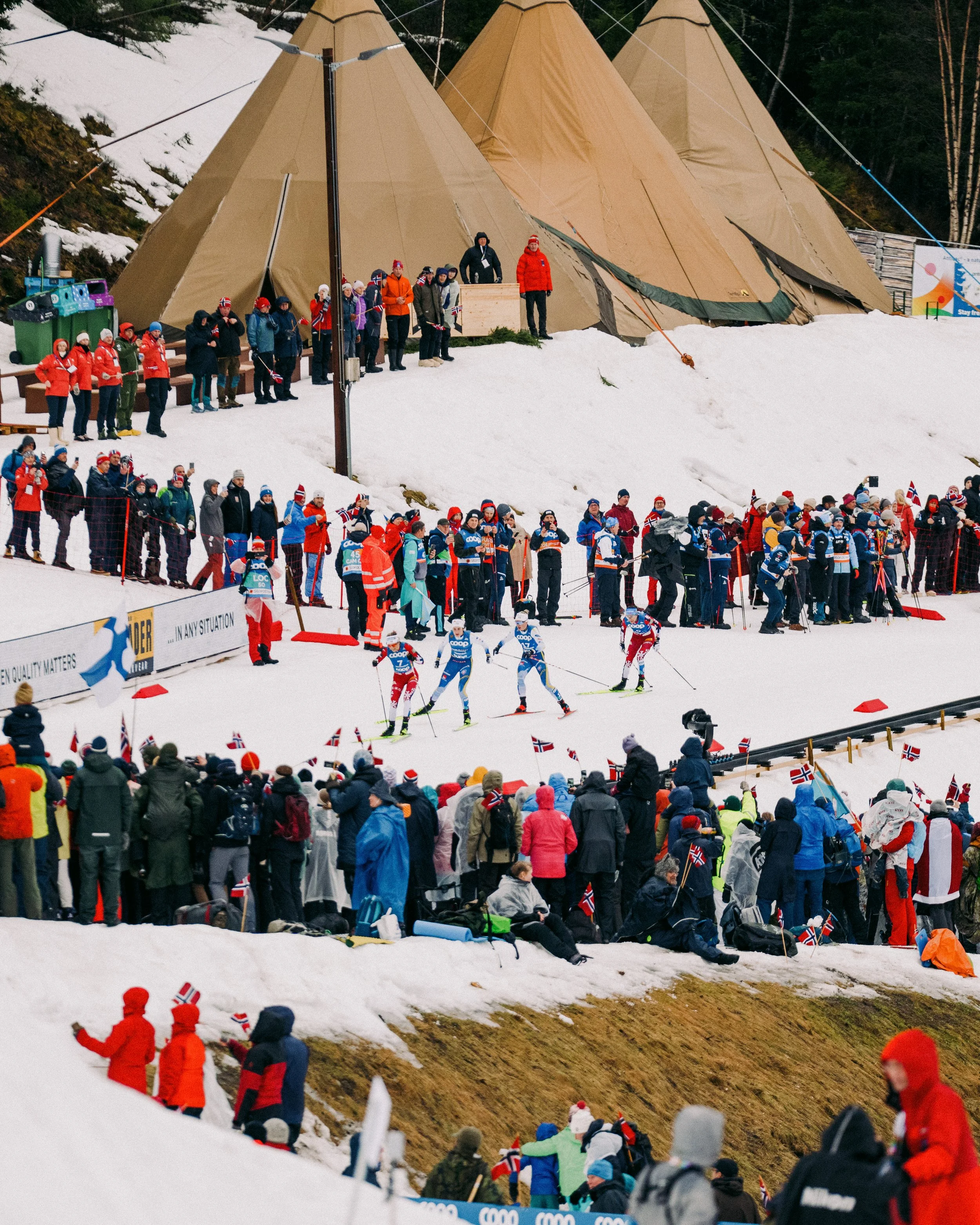 Trondheim 2025 crowd cheering at a cross-country ski race on snow-covered terrain, with athletes in racing suits and Norwegian flags visible.