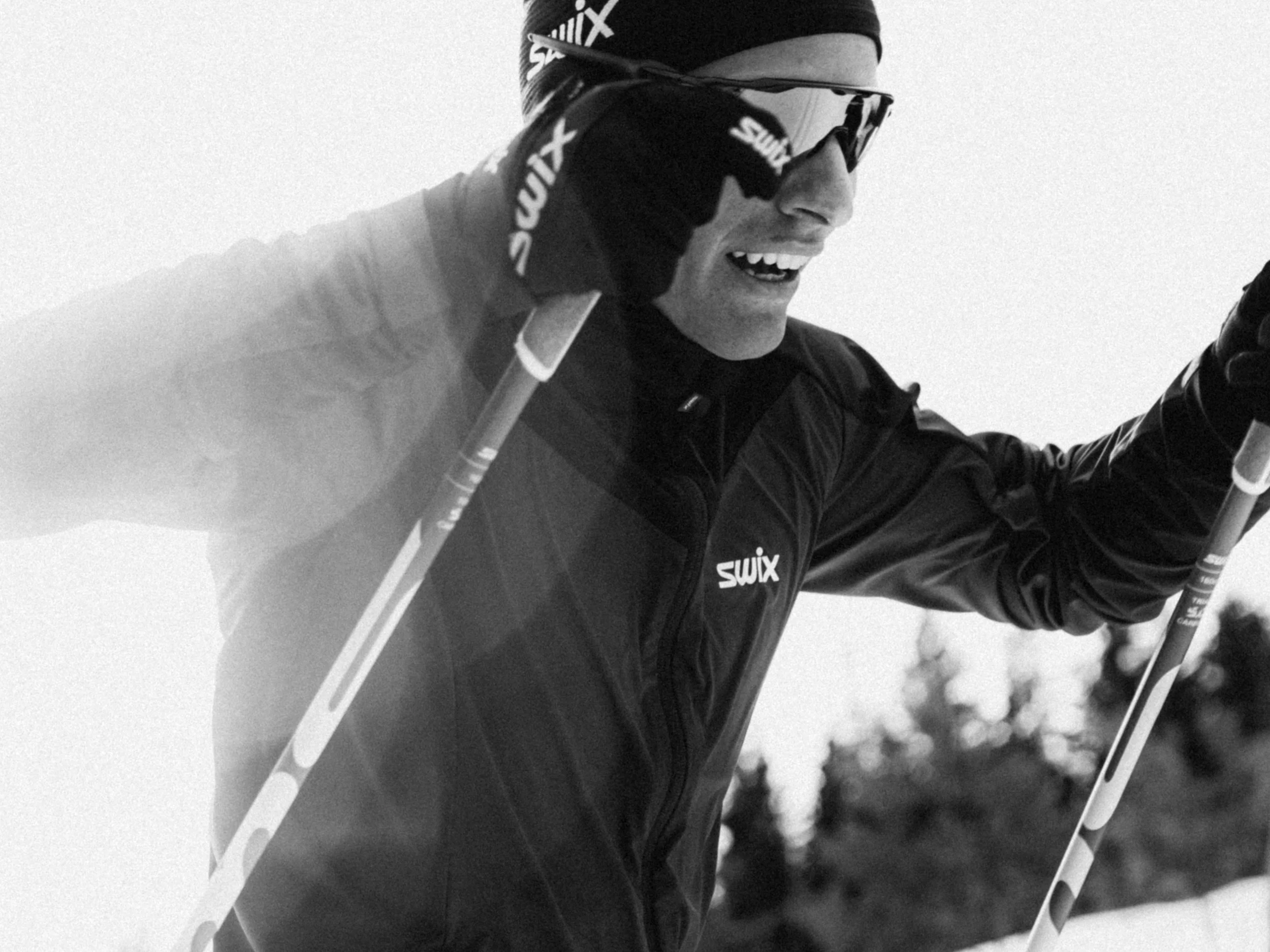 A man in winter sports gear, including a SWIX branded jacket, sunglasses, and a headband, Crosses Ski poles while participating in a ski race or activity outdoors.