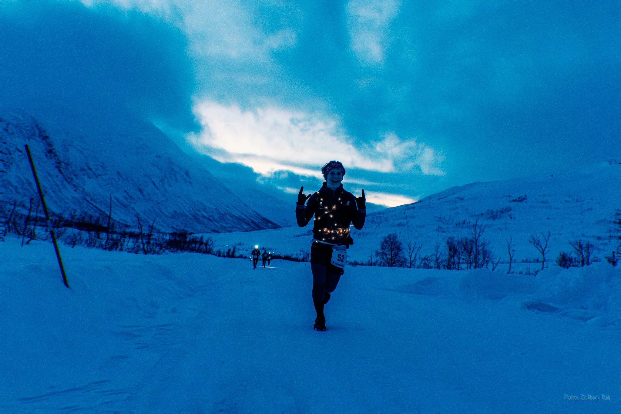 A person running in a snowy landscape during dusk or dawn, wearing a black outfit with Christmas lights and showing a happy gesture with both middle fingers raised.