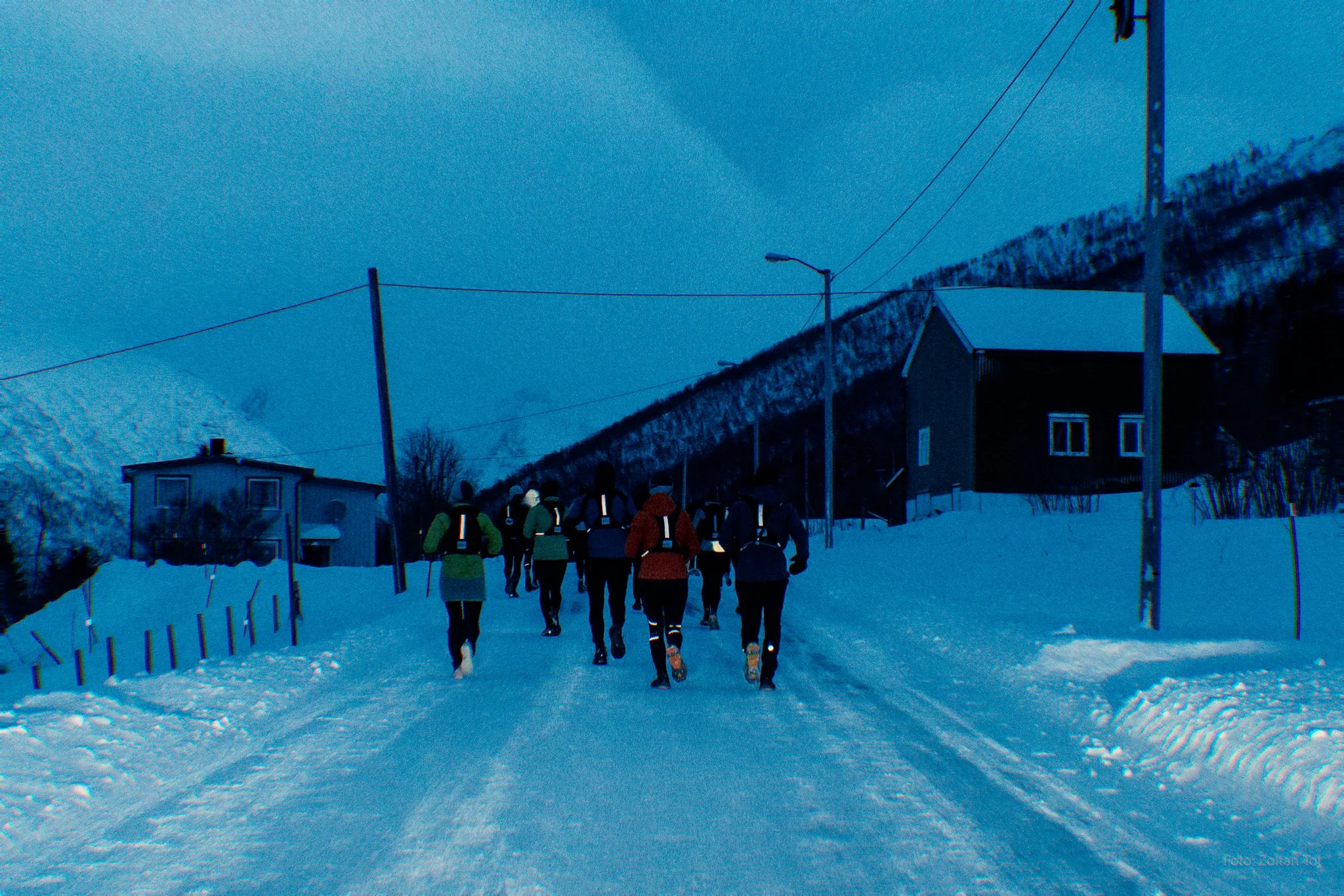 A group of people in winter clothing, running on a snow-covered road in a rural area with houses and snow-covered mountains in the background.