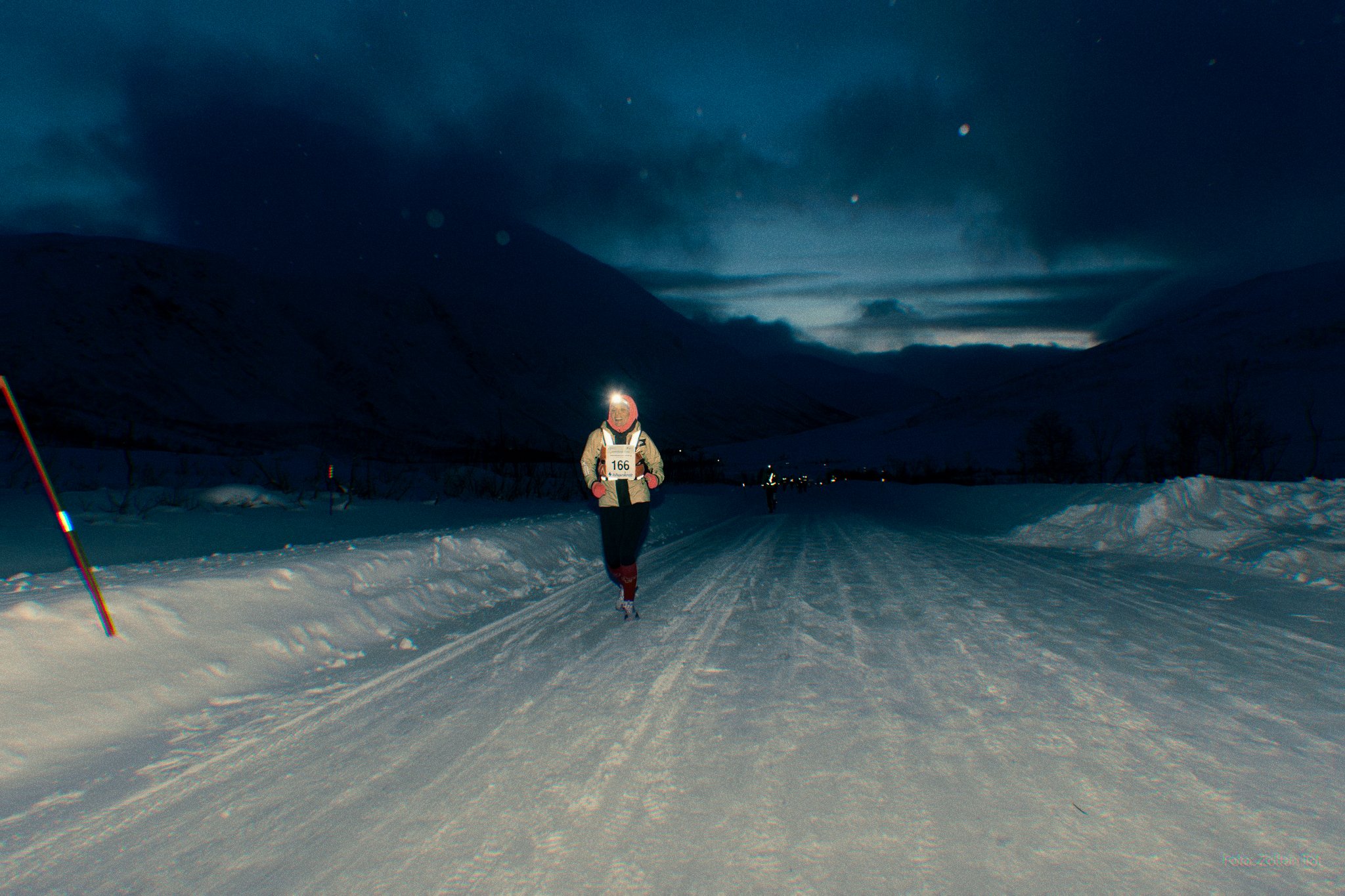 A person running on a snowy trail in a mountainous landscape at dusk or dawn, wearing winter clothing and a headlamp, with mountains in the background and a cloudy sky overhead.