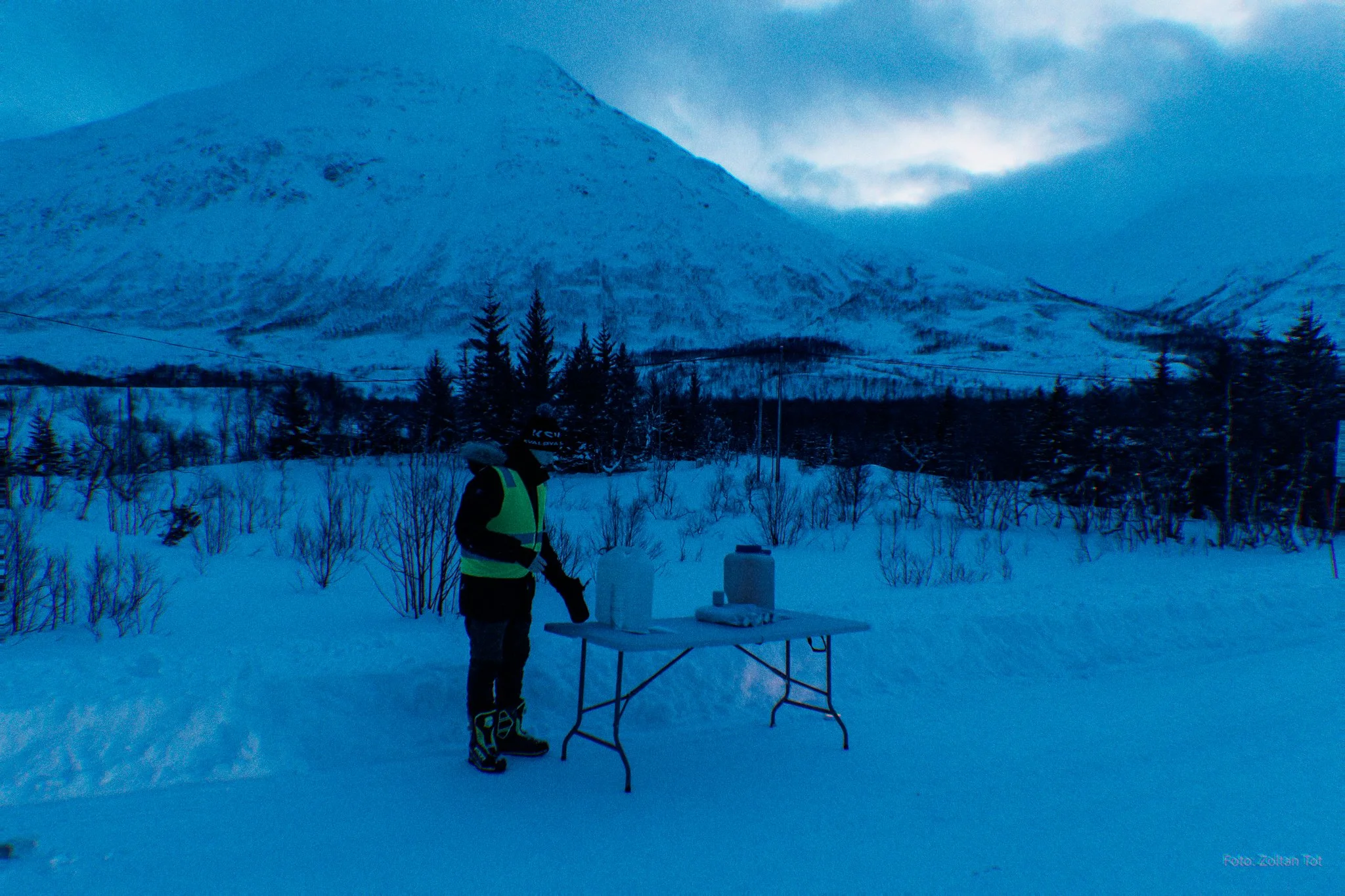 A person in winter gear and a reflective vest standing at a table with containers on snow, with snow-covered mountains and trees in the background during twilight.