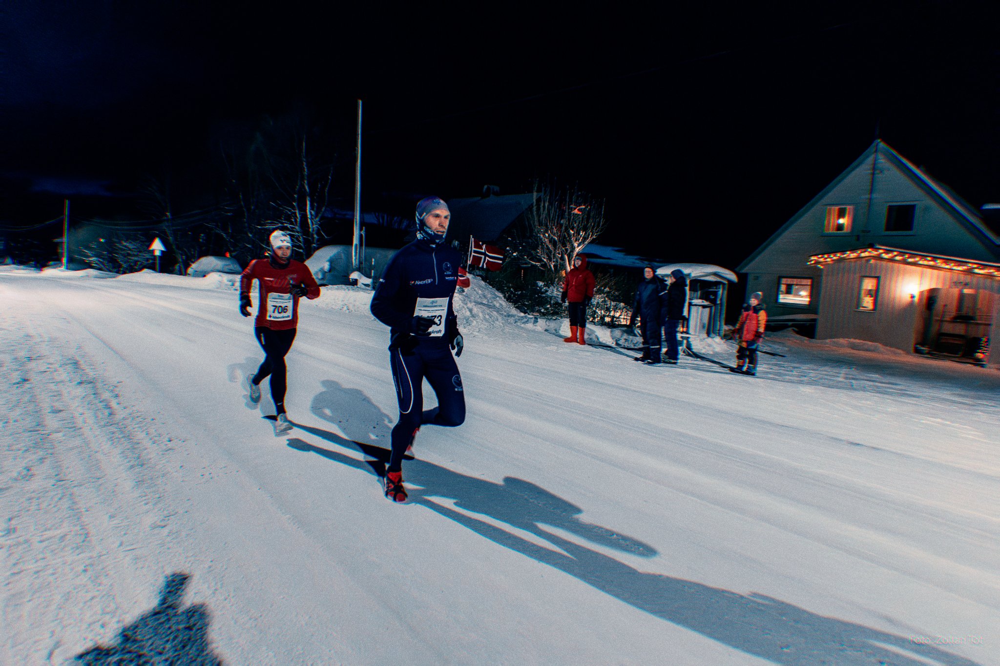 Nighttime view of three people running in a snow-covered street, dressed in winter athletic clothing, with spectators and houses decorated with Christmas lights in the background.