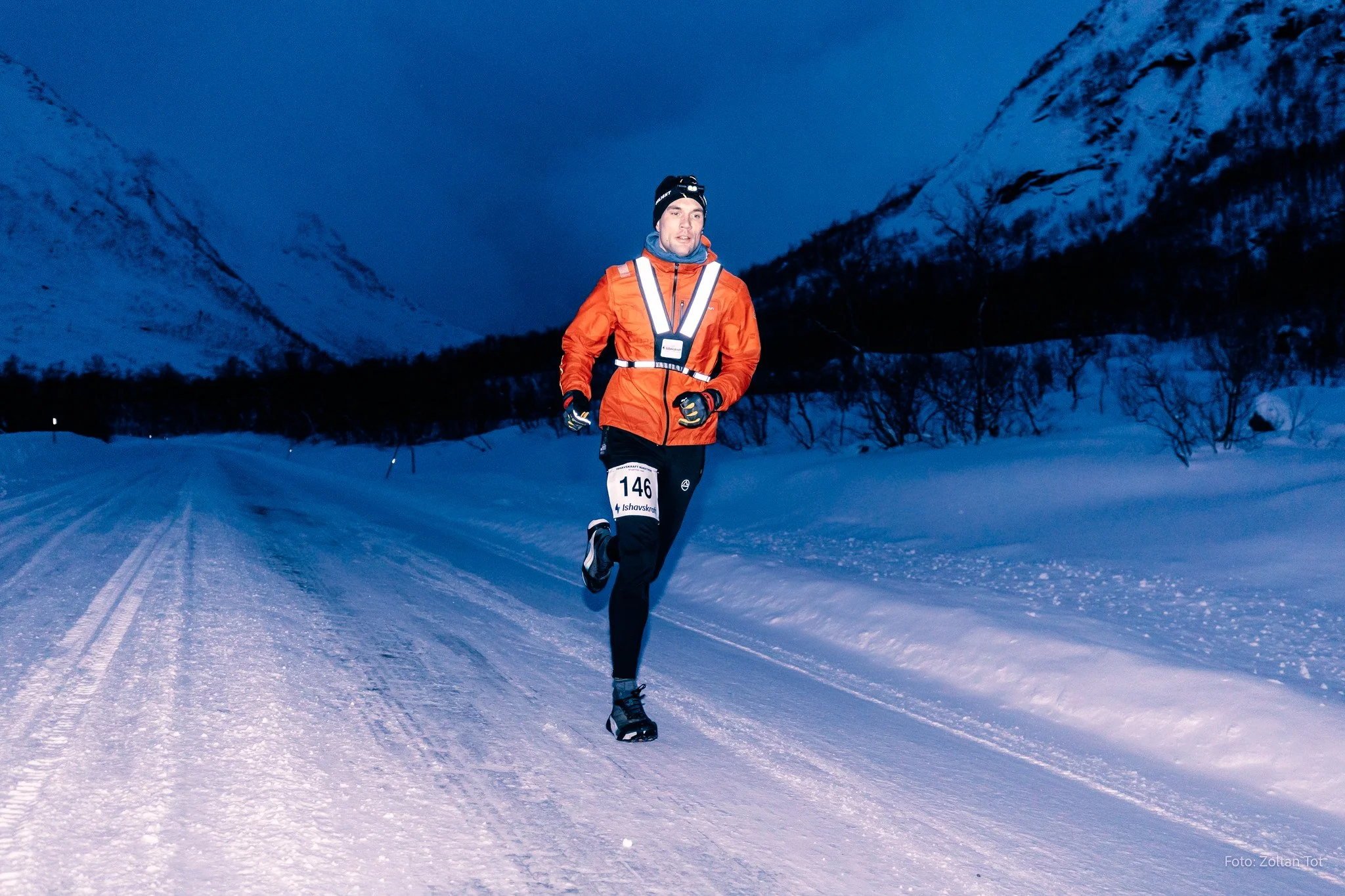 A man running on a snow-covered trail in a mountainous winter landscape during twilight, wearing an orange jacket, black pants, and a headlamp.