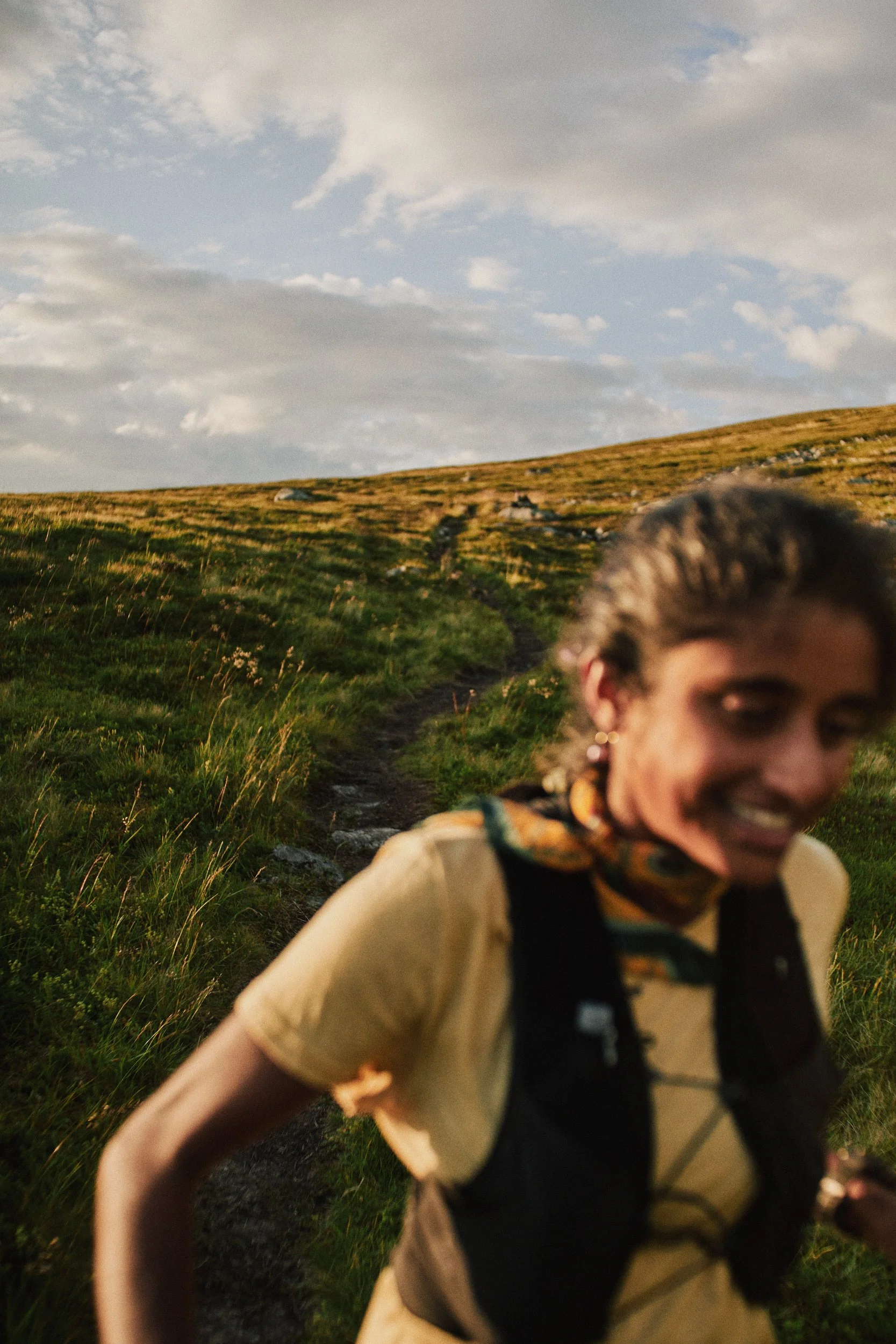 Shanga Ba with a backpack and scarf on a hiking trail in a grassy hillside landscape with cloudy sky.