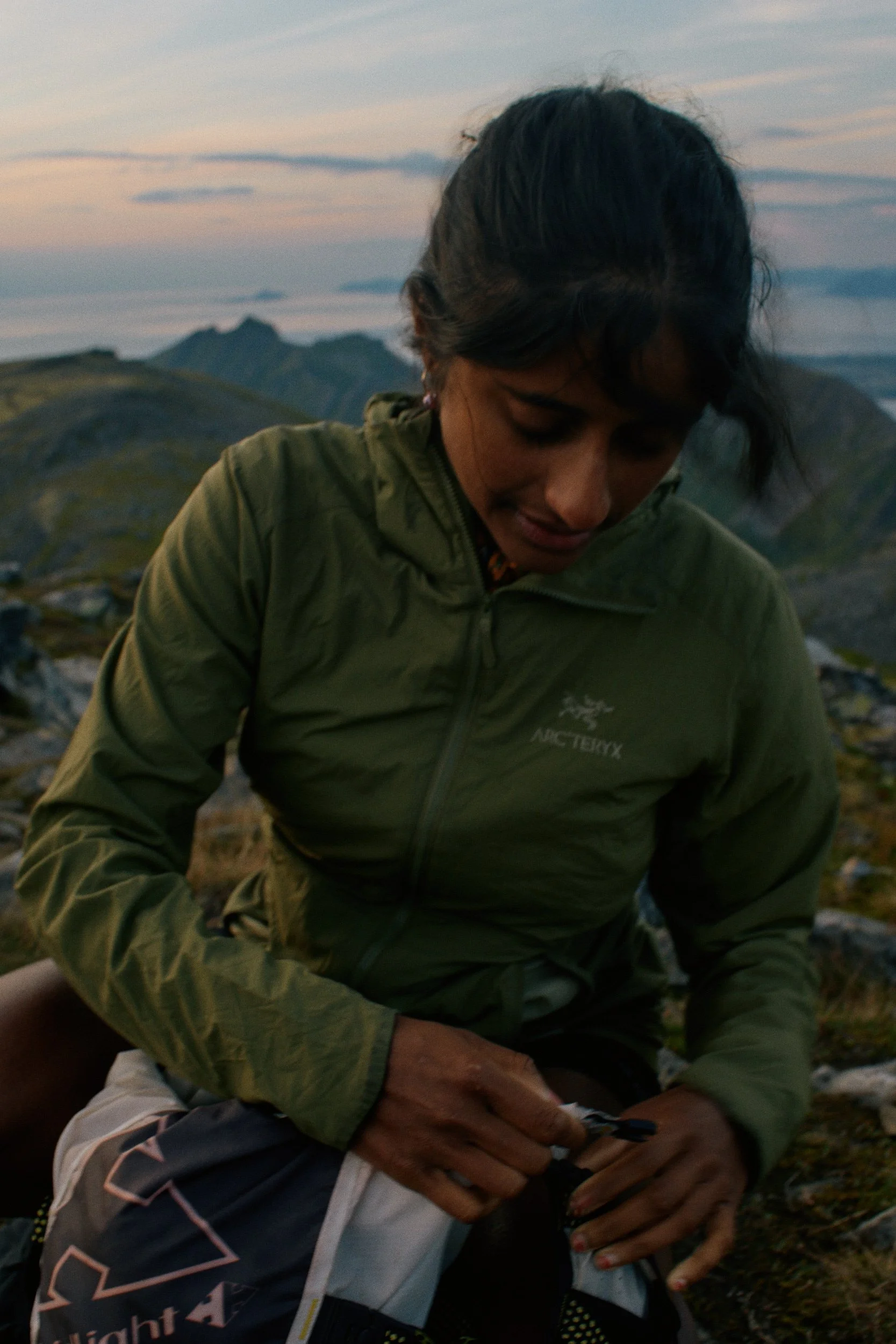Shanga Ba in a green jacket outdoors during sunset, with mountains in the background, adjusting her gear.