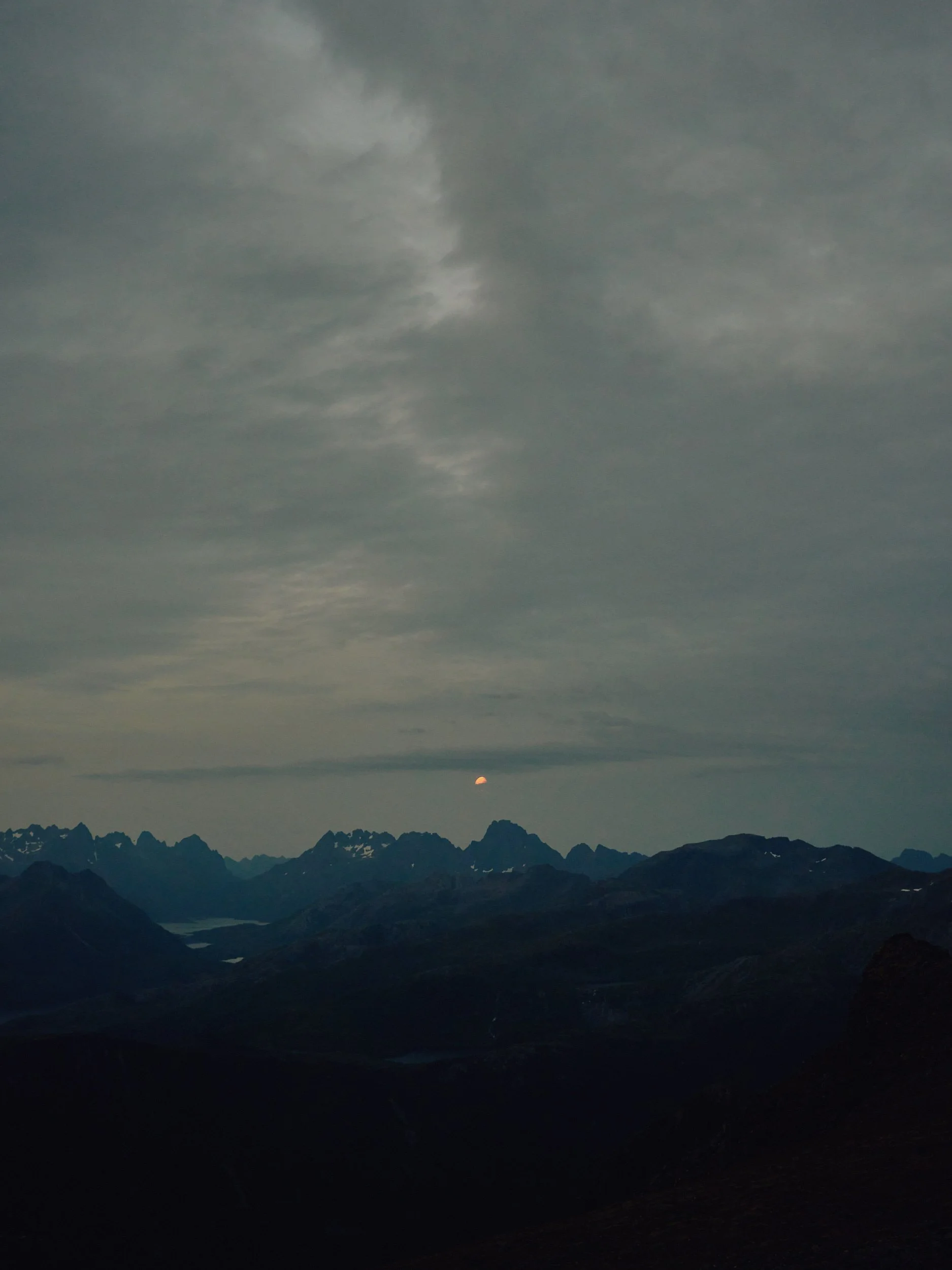 Mountain landscape at dusk with a small orange moon partially out of view behind clouds and a range of jagged mountains.