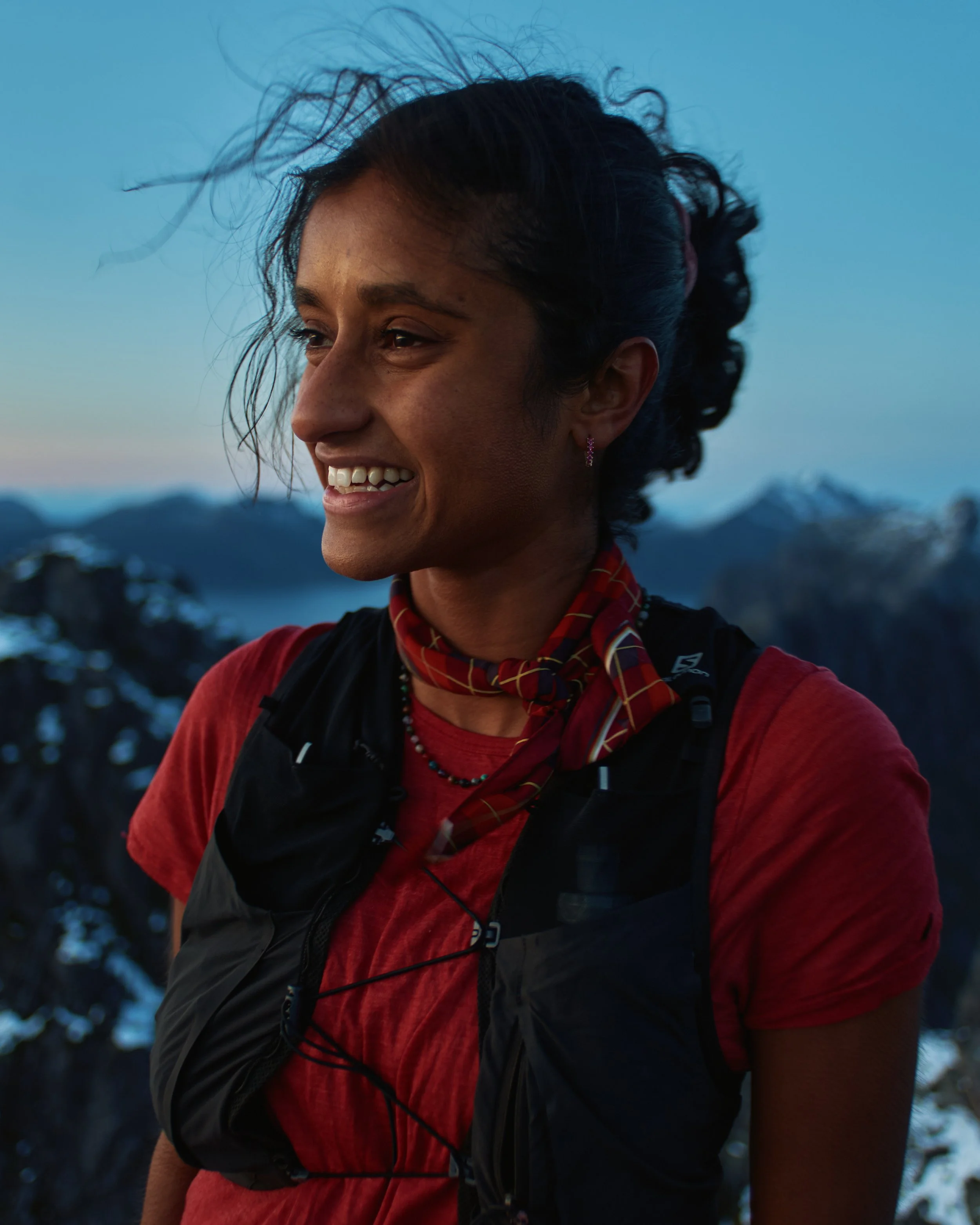 Shanga Ba smiling in outdoor mountain setting during dusk, wearing a red shirt, black vest, colorful scarf around her neck, and a hydration backpack.