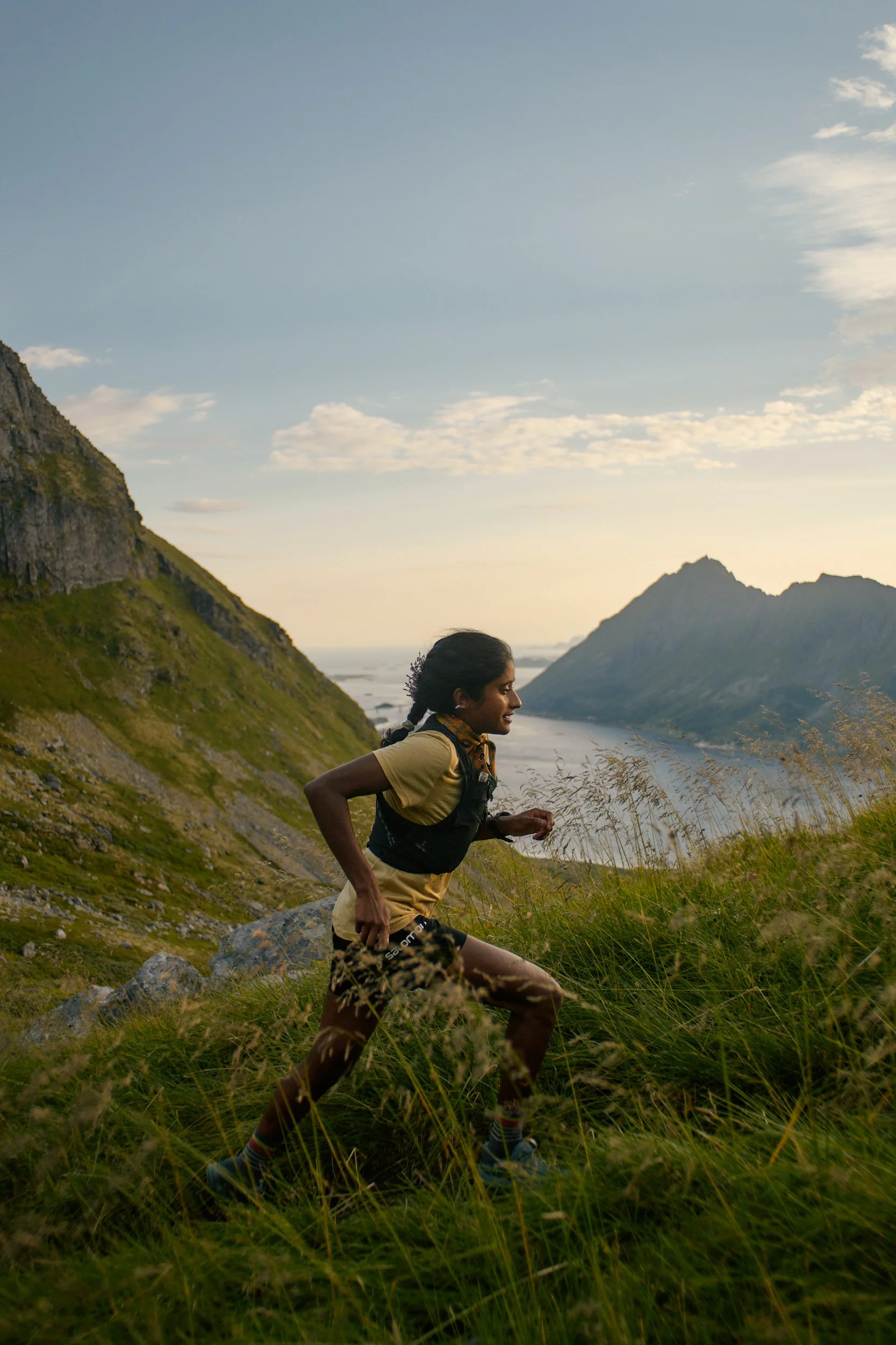 A woman trail running on a grassy hillside with mountains and water in the background during sunset.