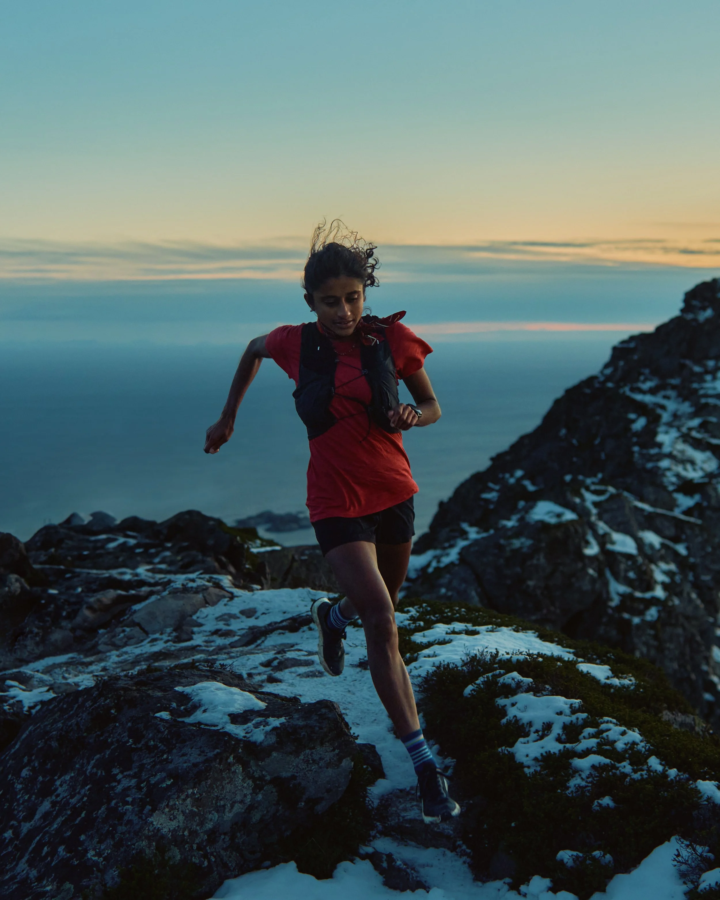 A woman running on snowy rocks near a mountain with a sunset sky in the background.
